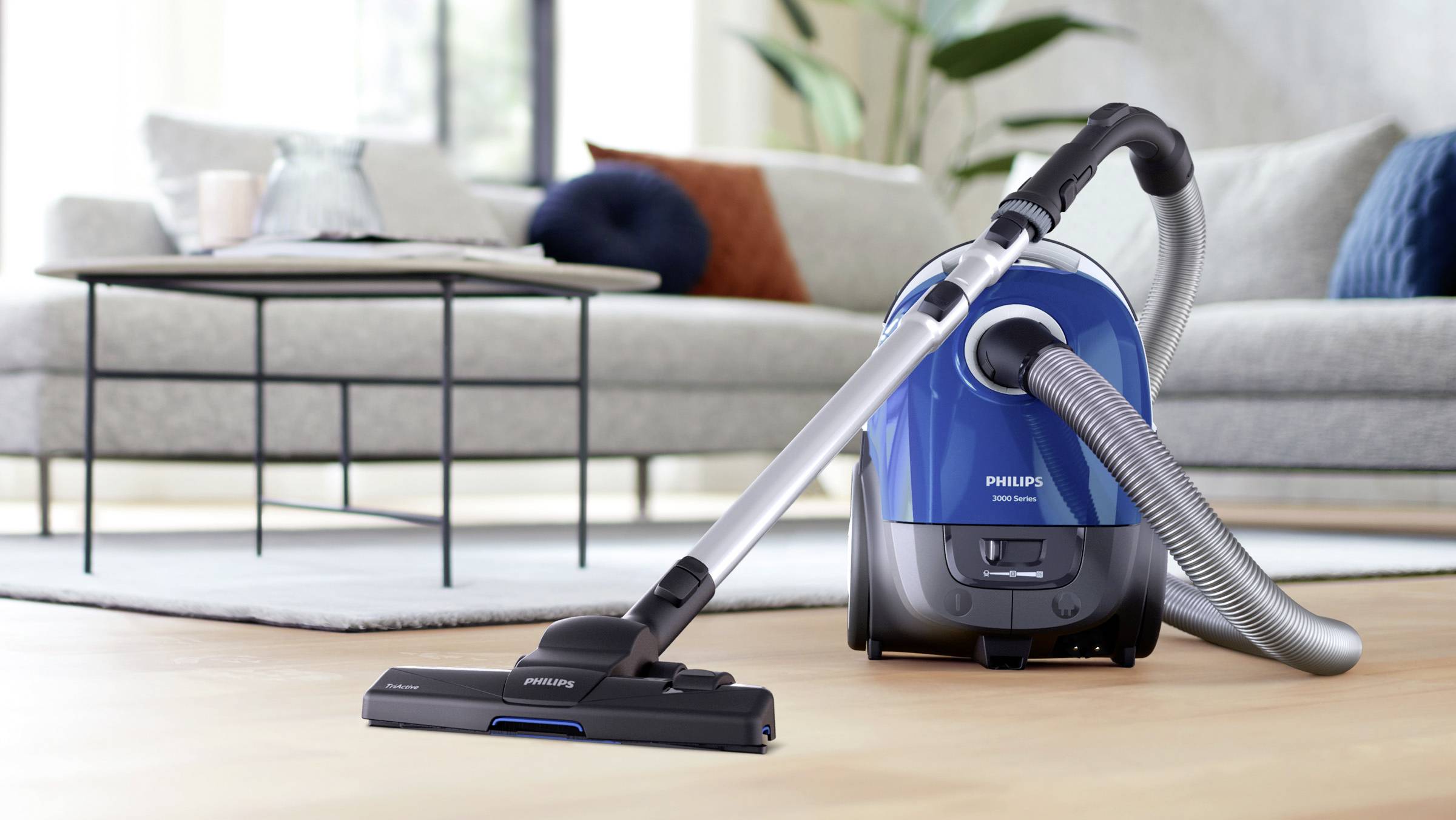 Un aspirateur bleu est allumé et posé sur un plancher en bois dans un salon avec un canapé gris et une table basse en arrière-plan.