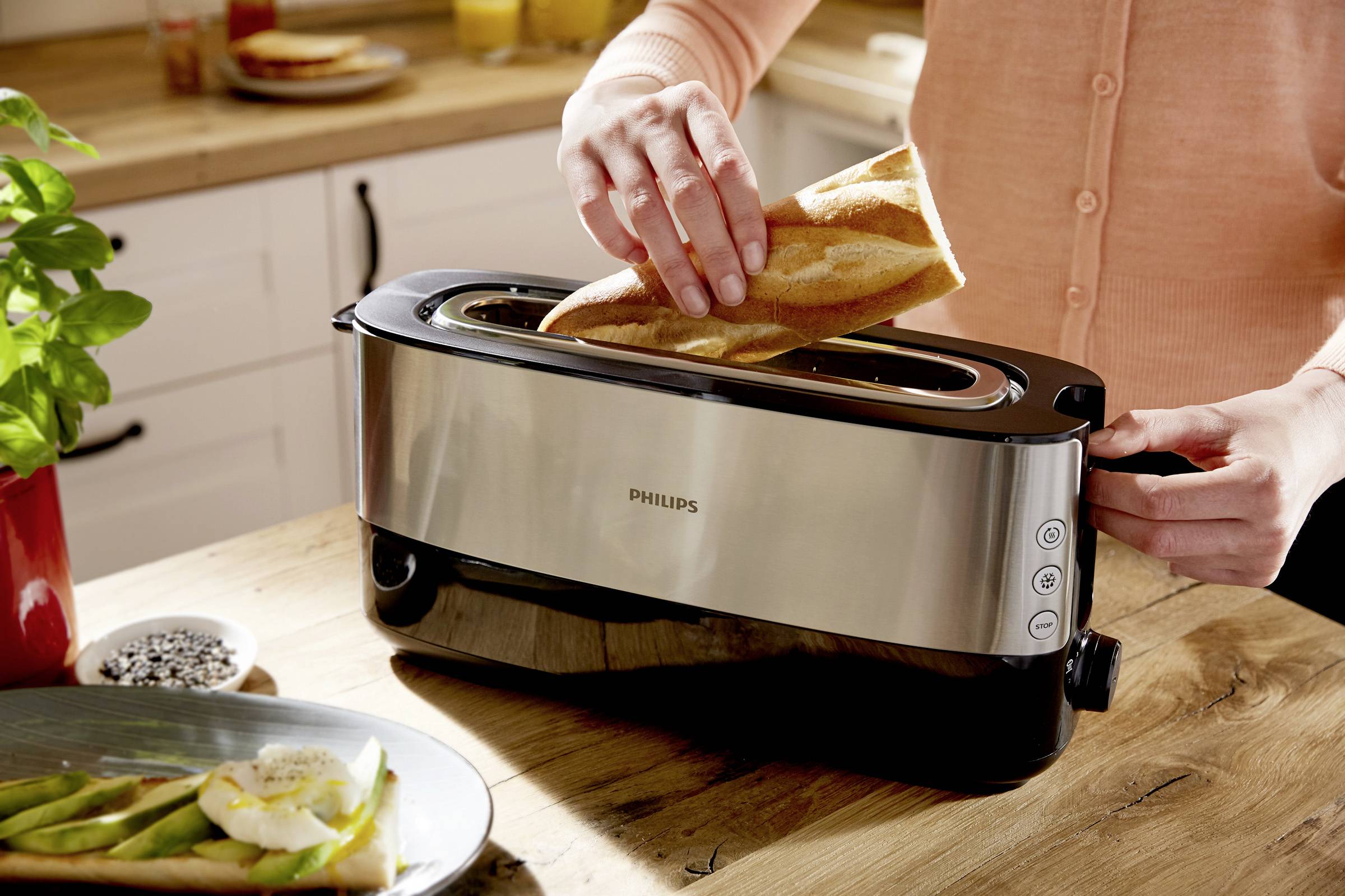 Une personne insère un morceau de baguette dans un grille-pain long et noir posé sur un plan de travail de cuisine. À côté, des assiettes avec des tartines.