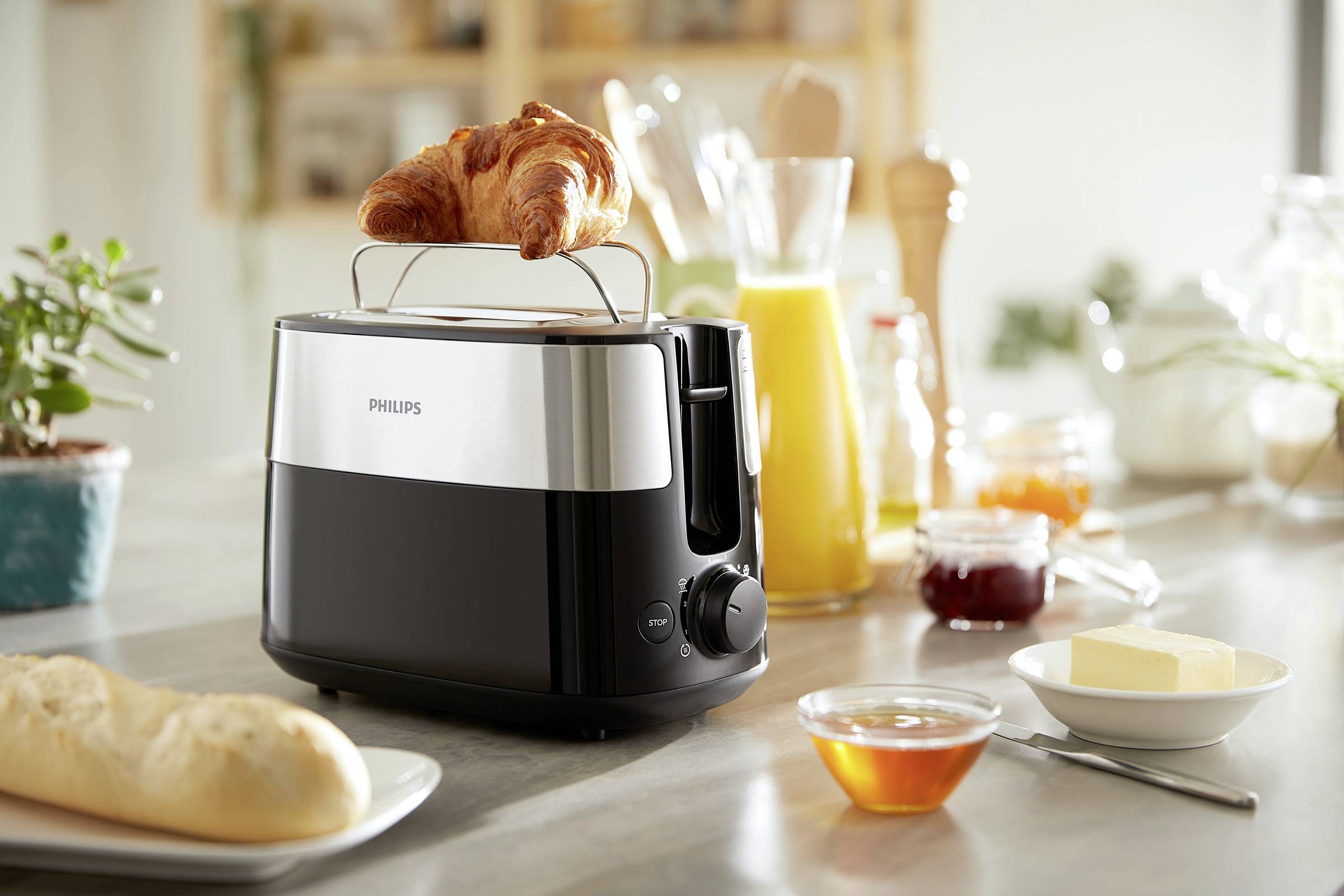 Un grille-pain avec un croissant posé dessus est placé sur une table, entouré de pain, de beurre, de confiture et d'un verre de jus d'orange dans une cuisine.