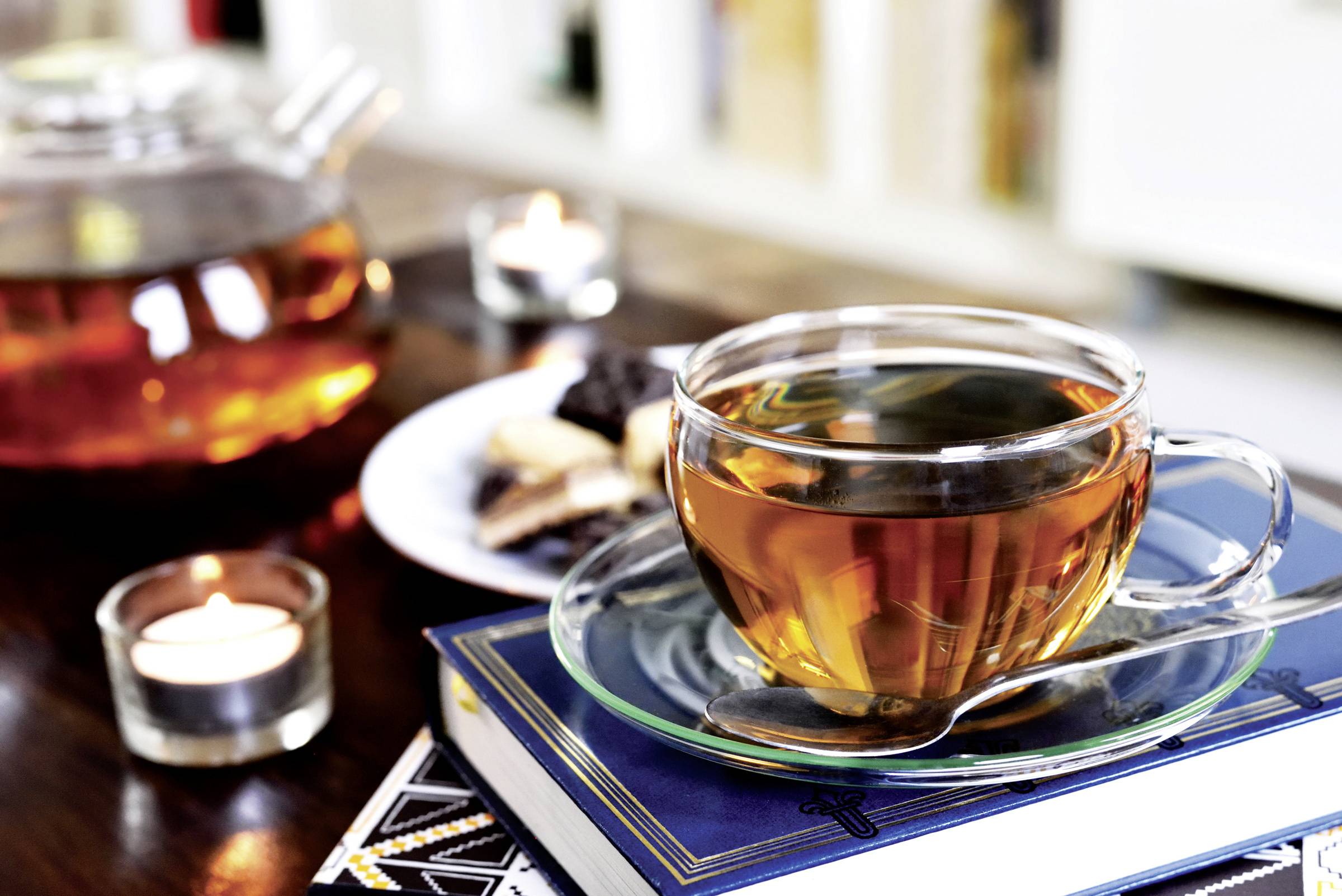 Une tasse de thé posée sur une pile de livres, à côté de bougies et d'un plateau de pâtisseries, illustrant une ambiance calme et chaleureuse.