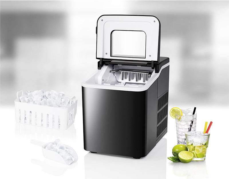 Préparateur de glaçons noir avec couvercle ouvert à côté d'un panier rempli de glaçons, d'une cuillère à mesurer et de boissons avec des citrons verts et des citrons.