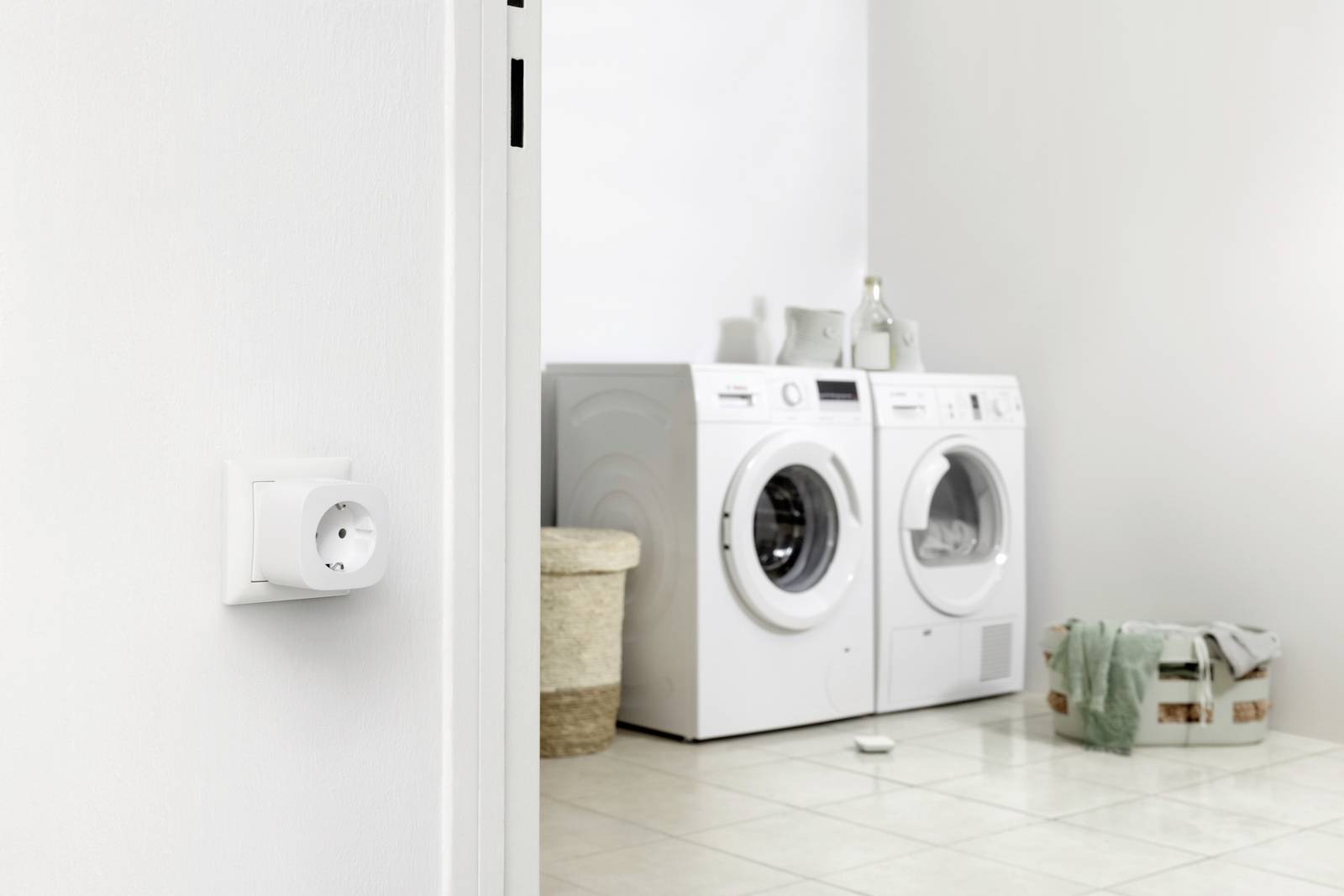 Buanderie avec un lave-linge et un sèche-linge côte à côte. Un panier à linge se trouve à droite, un autre avec un couvercle est placé à côté à gauche. Une prise électrique est accrochée au mur.