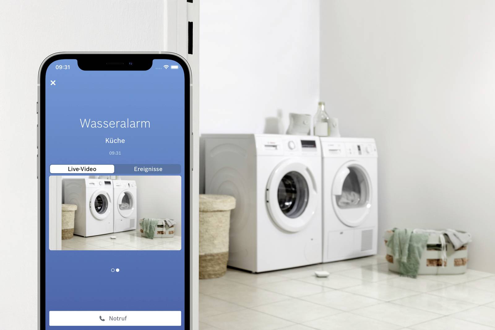 Un smartphone affiche une "alerte inondation" dans la cuisine. En arrière-plan, un lave-linge et un sèche-linge sont entourés de paniers à linge.