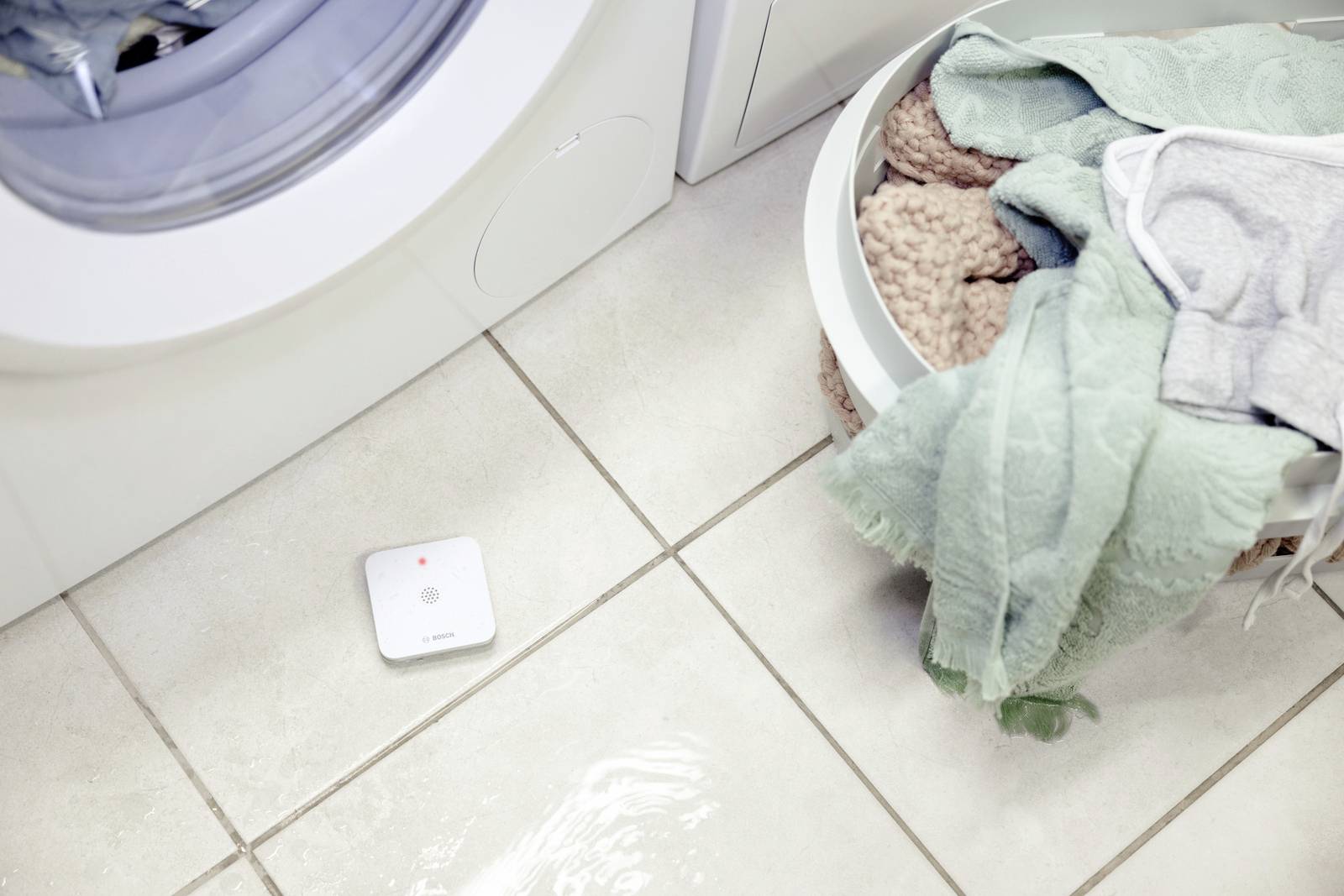 Un détecteur d'eau posé sur un sol carrelé à côté d'une machine à laver et d'un panier de linge. Le détecteur affiche un signal lumineux rouge.