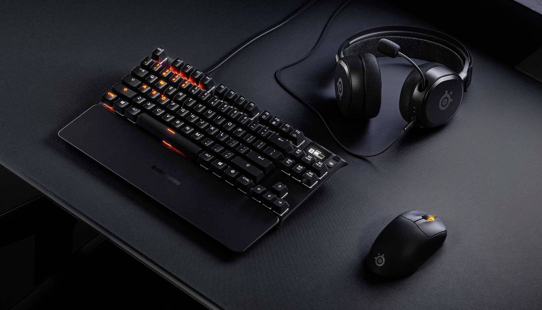 Clavier d'ordinateur avec des lumières orange, à côté d'une souris et d'un casque audio sur un bureau sombre.