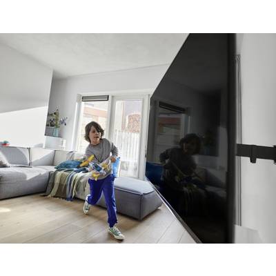 Un enfant joue joyeusement avec un jouet dans le salon, portant un haut gris et un pantalon bleu, devant un grand téléviseur.