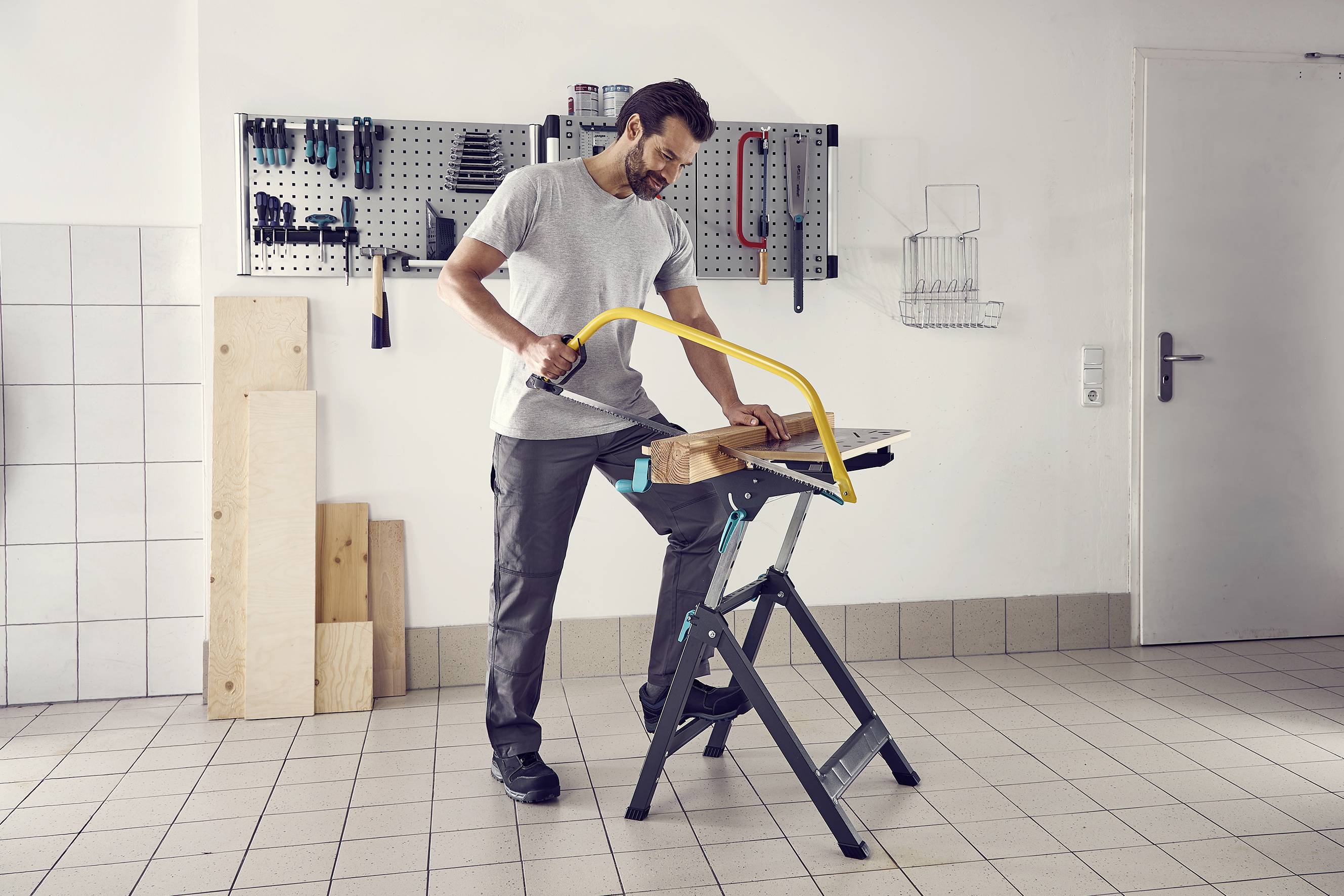 Un homme scie un morceau de bois avec une scie à main sur un établi dans un atelier. Des outils sont suspendus au mur en arrière-plan.