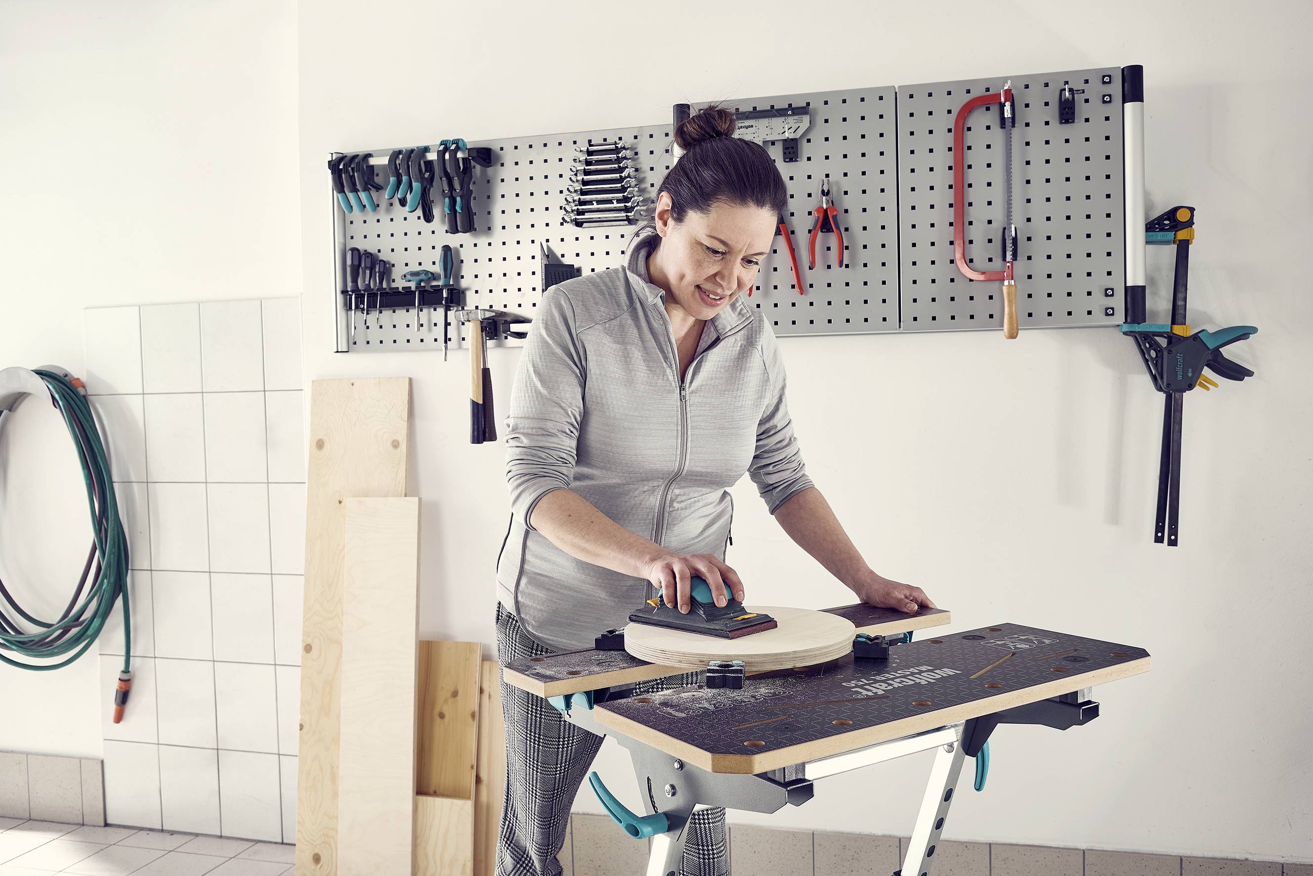 Une femme ponce un morceau de bois sur un établi. Des outils sont suspendus à un mur en arrière-plan.