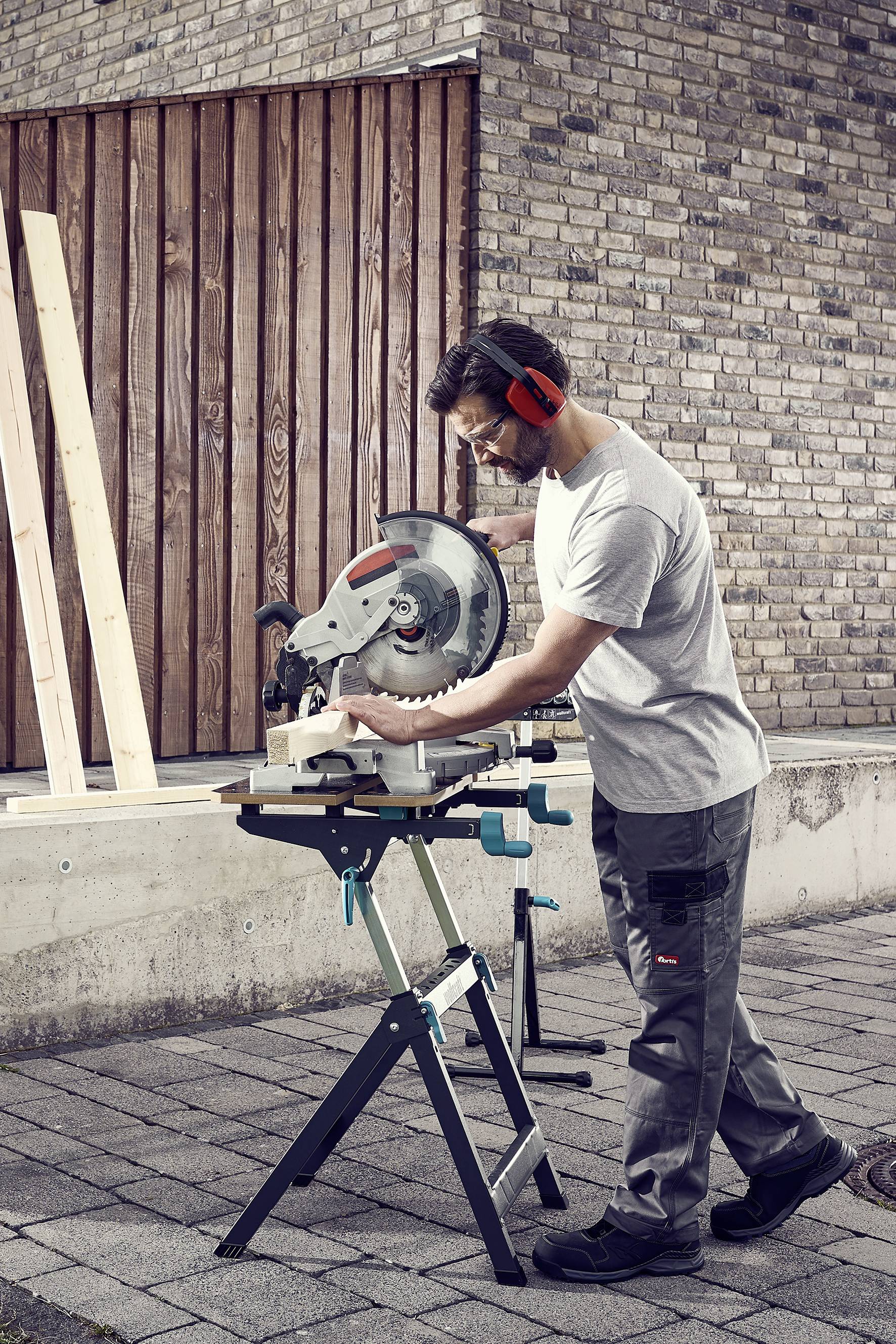 Un homme scie du bois avec une scie circulaire sur un support à l'extérieur. Il porte des lunettes de protection et des protections auditives.