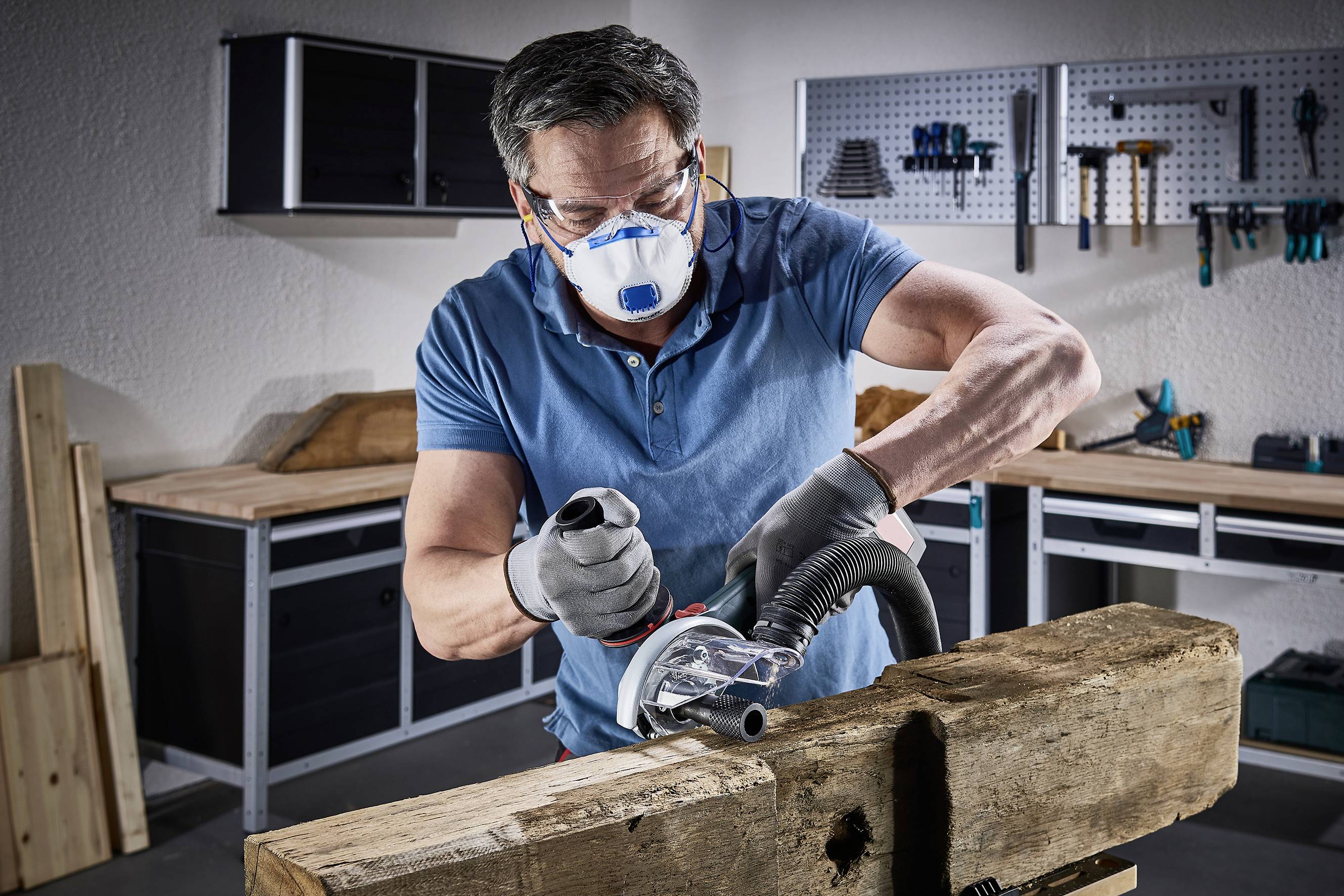 Un homme portant un masque de protection et des gants travaille dans un atelier avec une meuleuse d'angle sur un morceau de bois. Des outils sont visibles en arrière-plan.