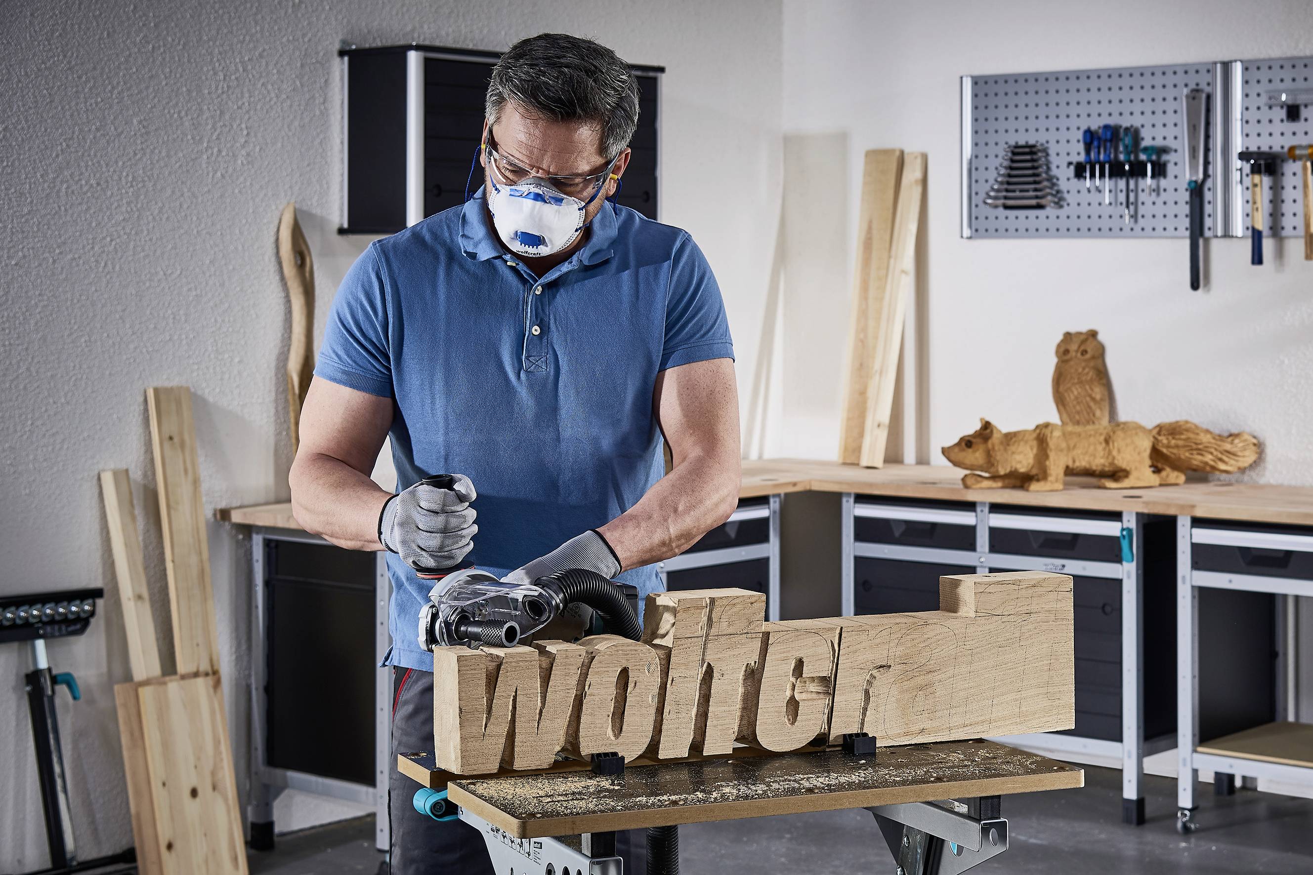 Un homme portant des lunettes de protection et un masque travaille dans un atelier. Il sculpte le nom 'Wolter' dans un morceau de bois. En arrière-plan : des étagères et des outils de travail du bois.