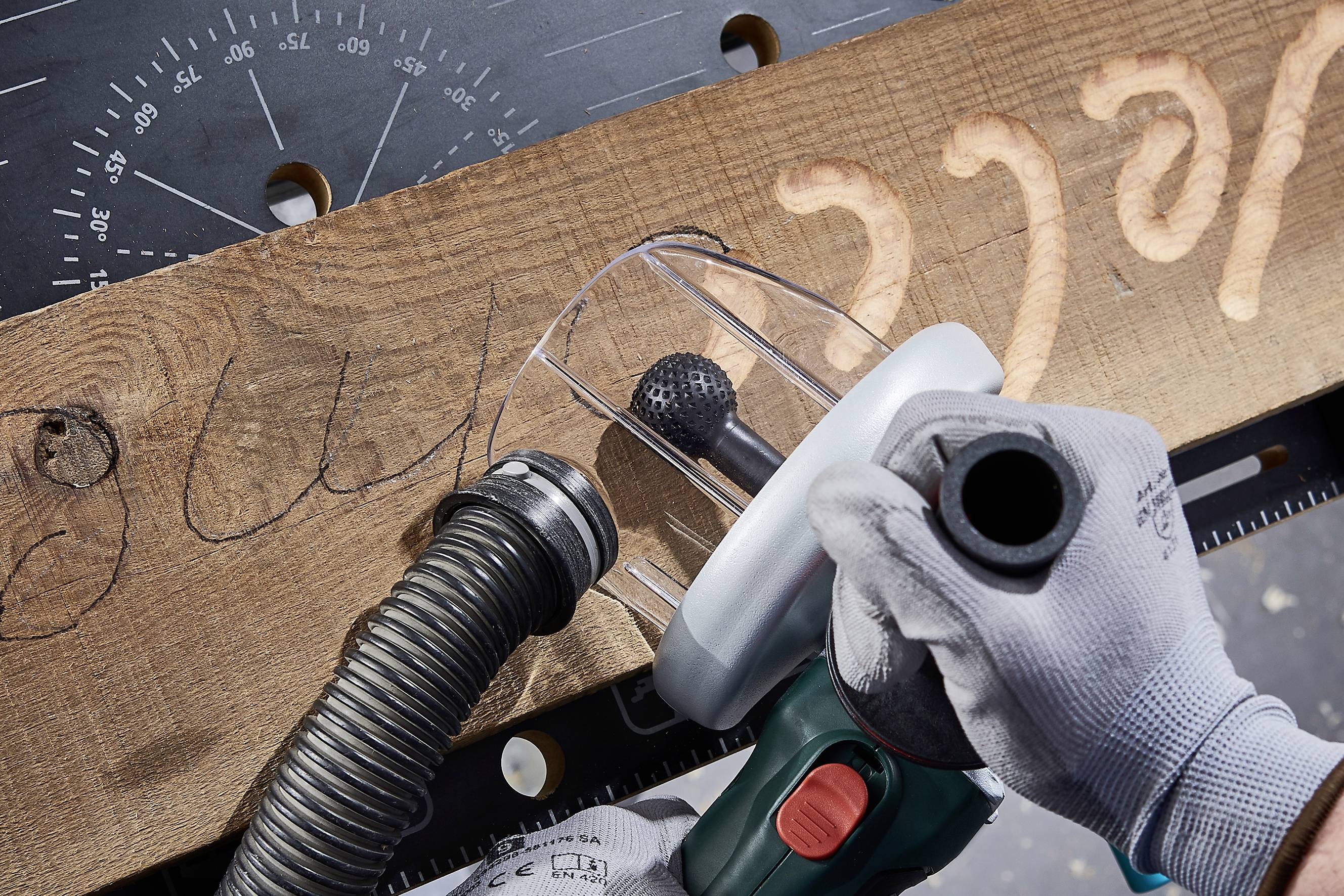 Une personne sculpte un motif décoratif sur une planche de bois à l'aide d'un outil électrique. Des dispositifs de protection et un tuyau d'aspiration sont visibles.
