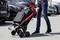 'Une personne portant un jean et des baskets pousse un caddie chargé sur un parking, des voitures en arrière-plan.'