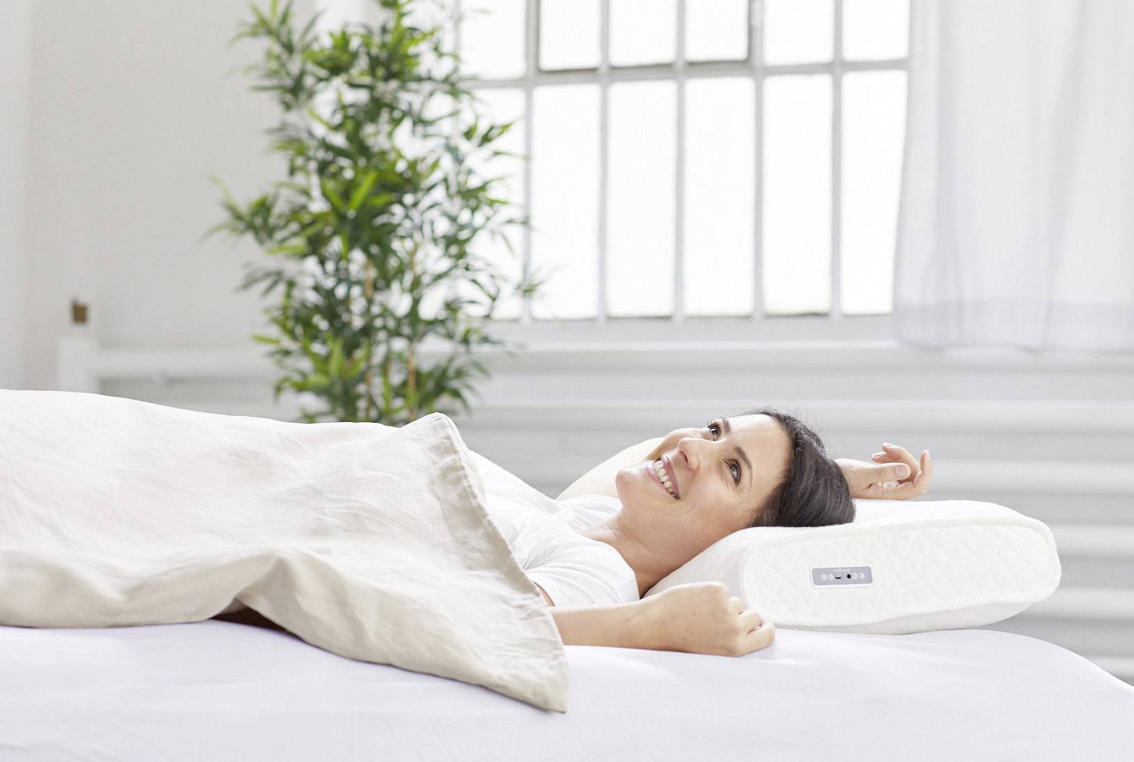Une femme est allongée de manière détendue sur un lit blanc, souriant, entourée d'un oreiller et de plantes en arrière-plan.