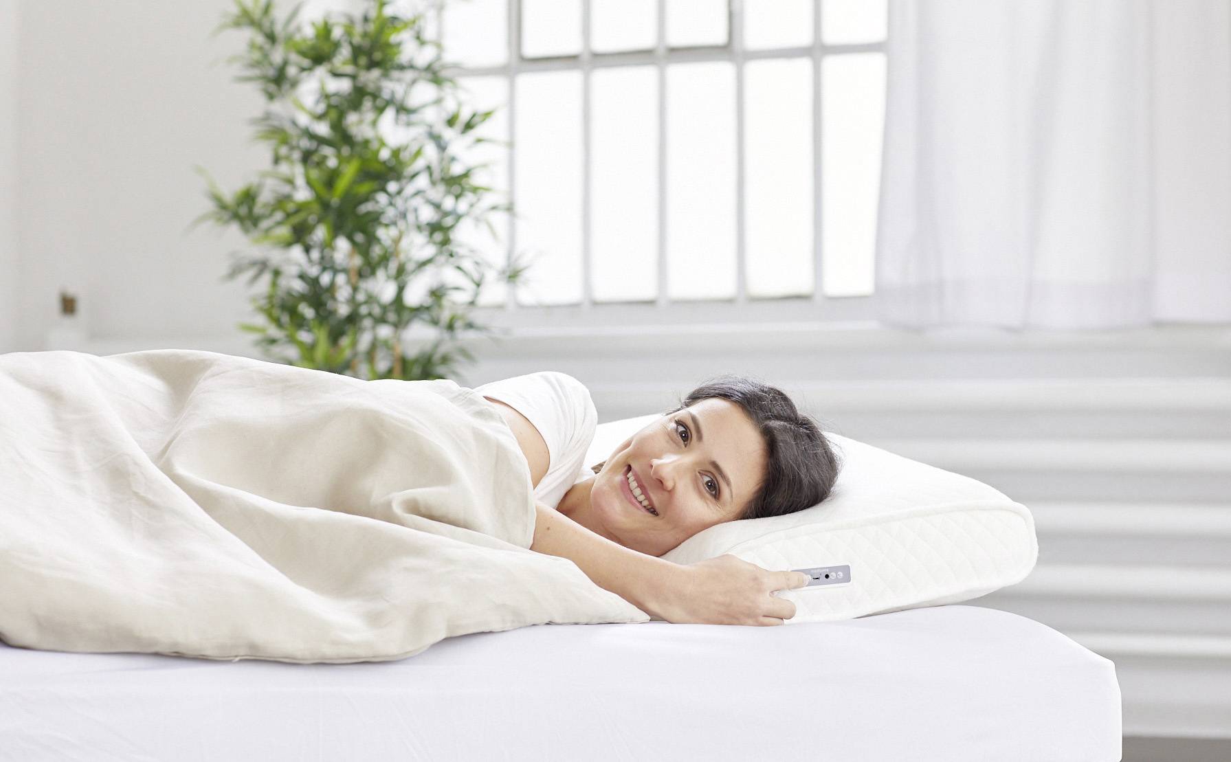 Une personne souriante dans un lit avec du linge de lit blanc, tenant un oreiller, dans une pièce lumineuse avec une plante en arrière-plan.