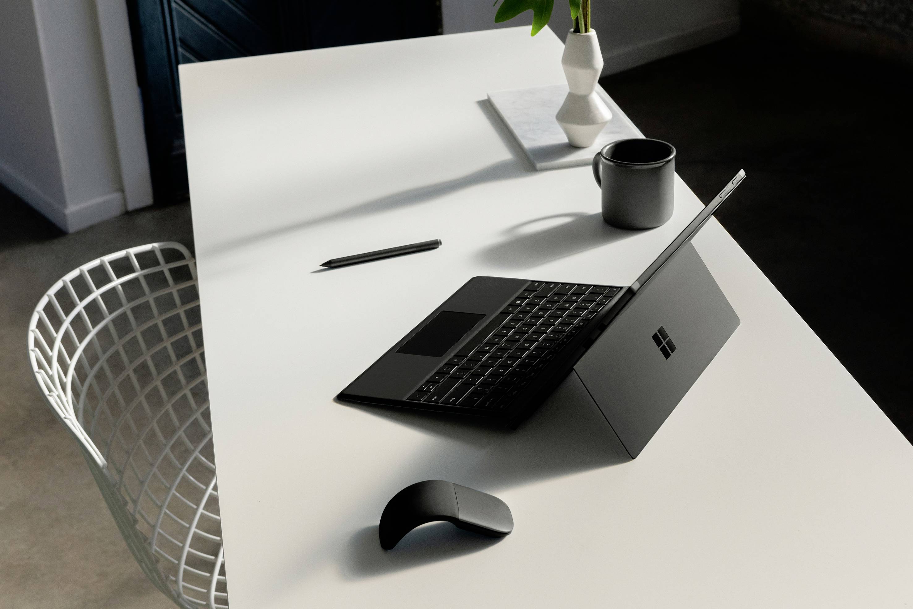 Un espace de travail moderne avec un ordinateur portable, une souris, un stylo, une tasse et un vase sur une table blanche à côté d'une chaise en métal.
