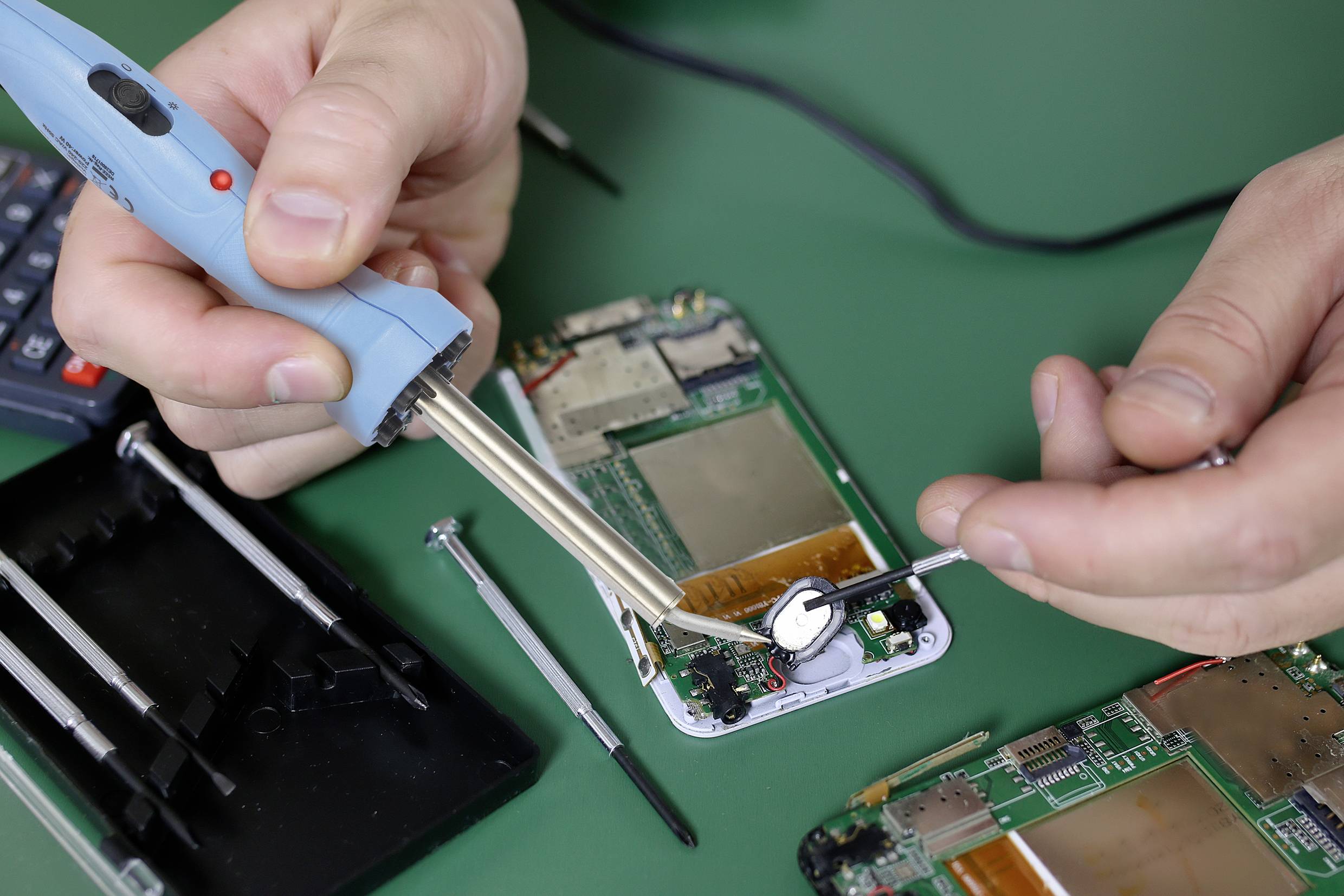 Une personne répare une carte électronique de téléphone portable ouverte à l'aide d'un fer à souder. Un tournevis et d'autres outils sont posés à côté.