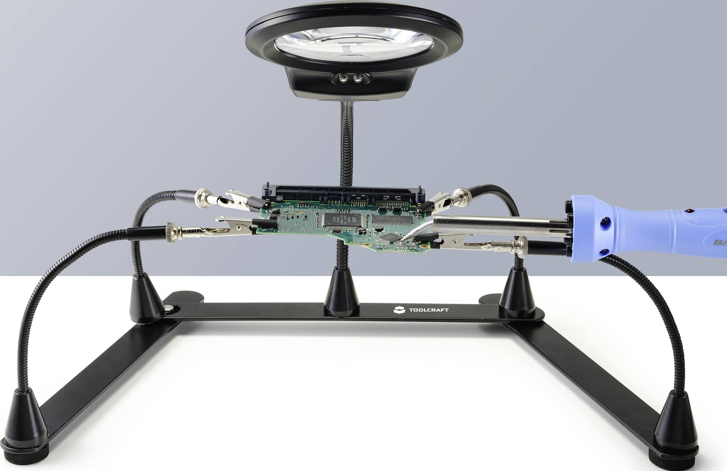 L'image montre un poste de soudage avec une carte électronique sous une loupe. Un fer à souder est prêt à intervenir, en contact avec la carte.