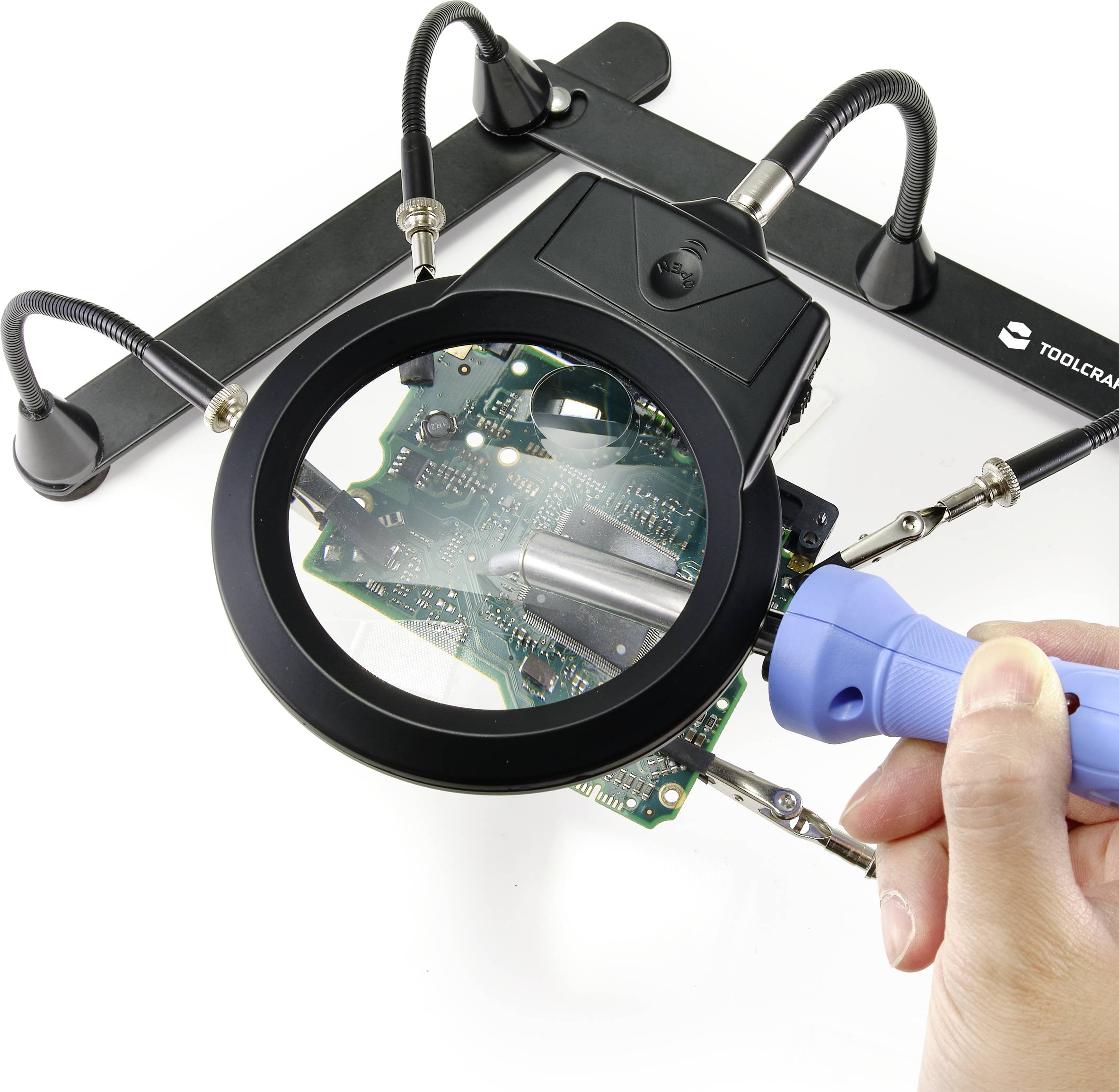 Une main tient un fer à souder bleu sous une loupe positionnée au-dessus d'une carte électronique.