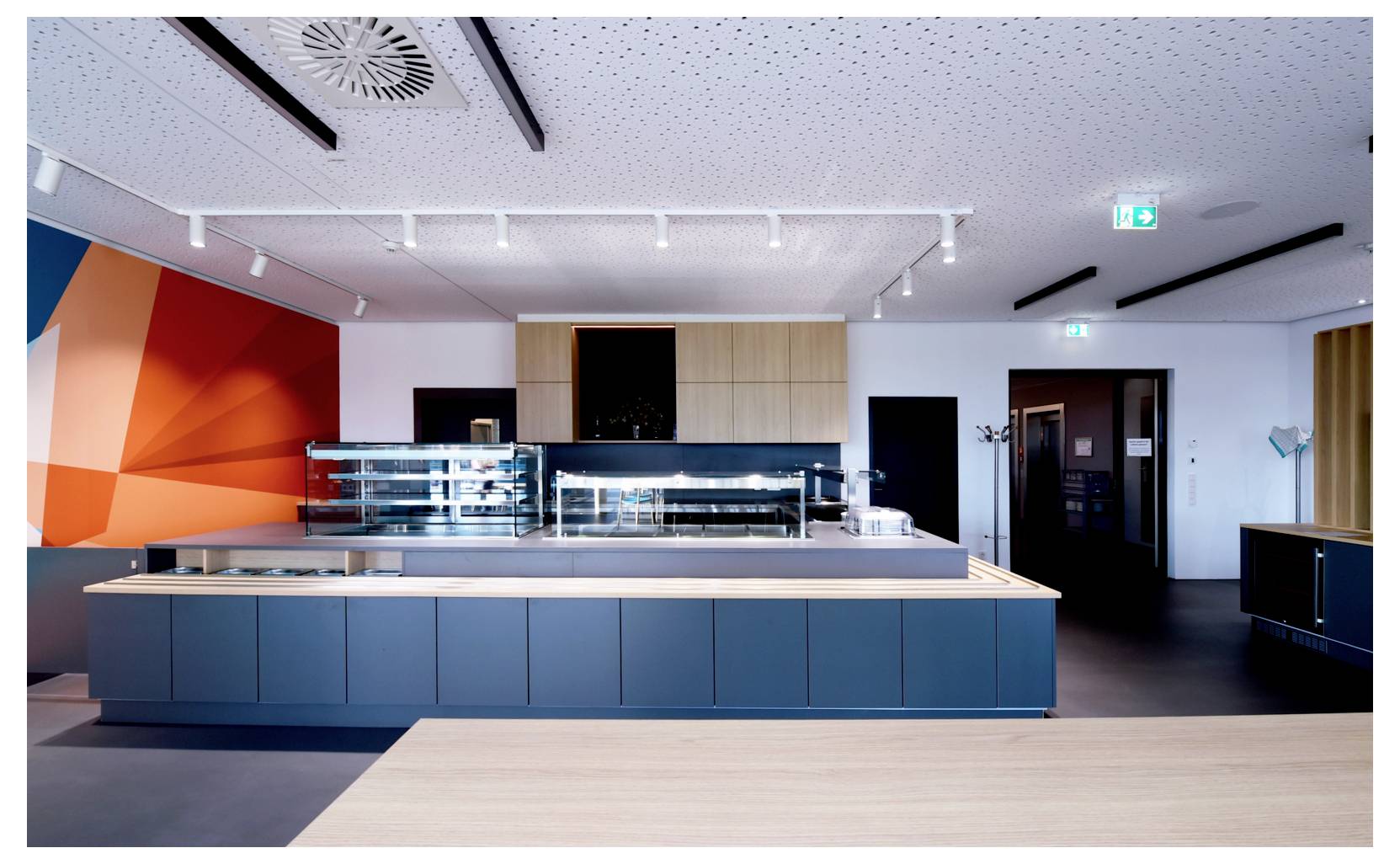 Intérieur moderne de cafétéria avec un grand comptoir de service central, des armoires bleues, des touches de bois et des œuvres murales géométriques orange sous des plafonniers lumineux.