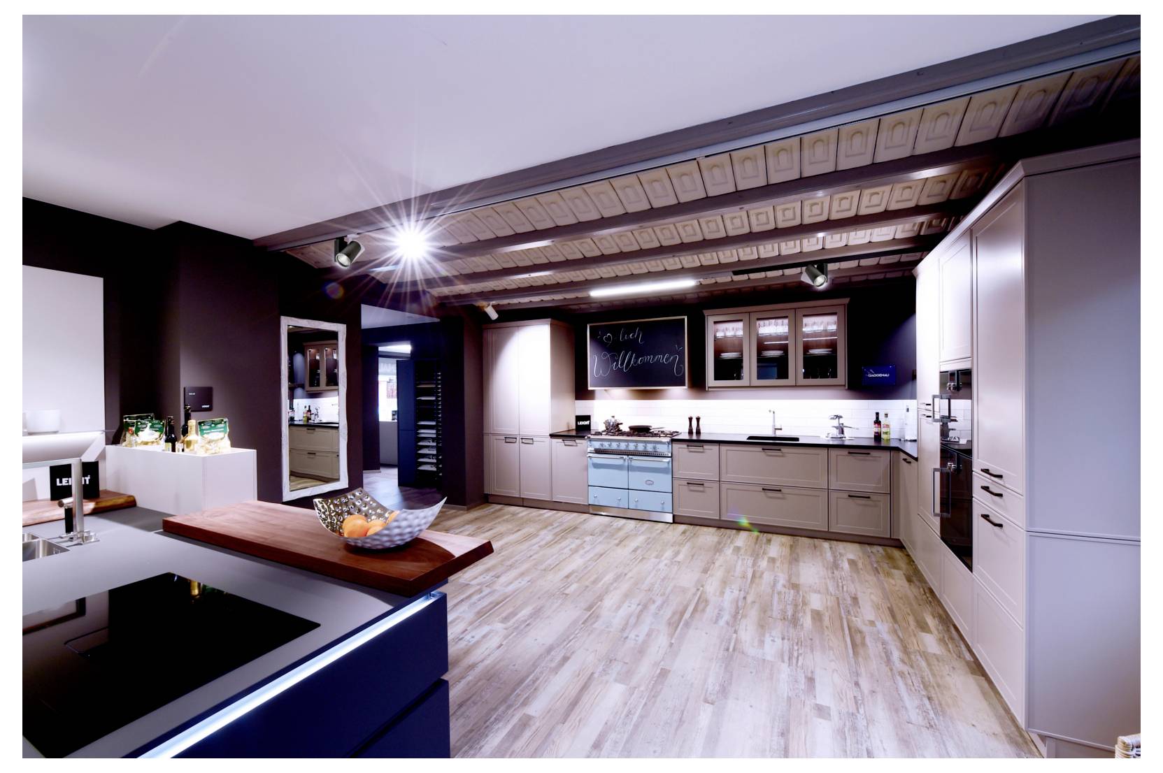 Salle d'exposition de cuisine moderne avec plafond à poutres apparentes, meubles clairs, îlot central, plaque de cuisson noire et électroménagers encastrés