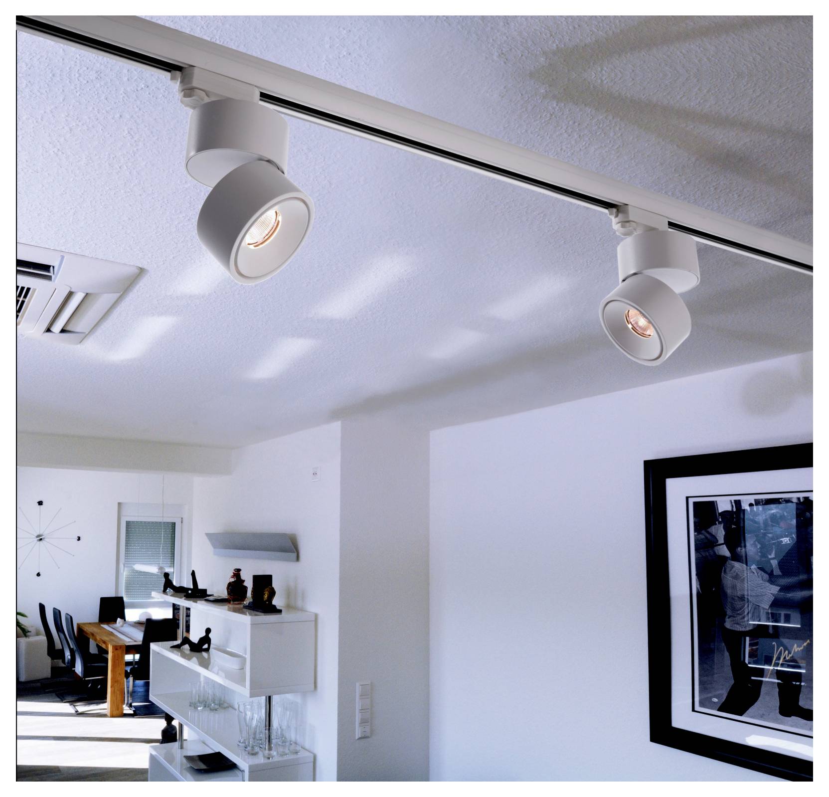 Pièce moderne avec des rails d'éclairage au plafond illuminant un intérieur blanc, avec une salle à manger et des tableaux muraux en arrière-plan.
