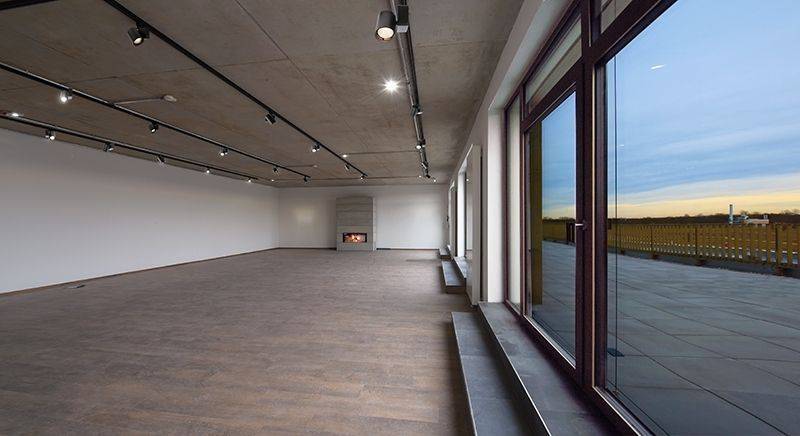 Un espace vide avec un sol en bois, de grandes fenêtres et une cheminée contre le mur. La vue extérieure montre une terrasse et un ciel immense.