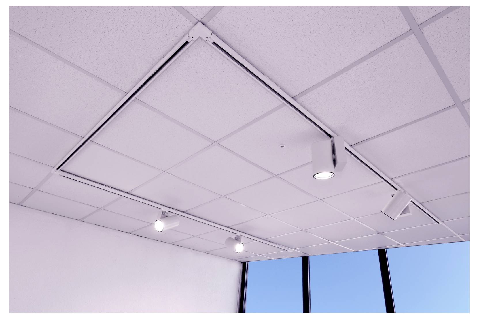 Plafond avec une trappe d'accès rectangulaire et des rails d'éclairage au-dessus d'une pièce dotée de grandes fenêtres teintées de bleu.
