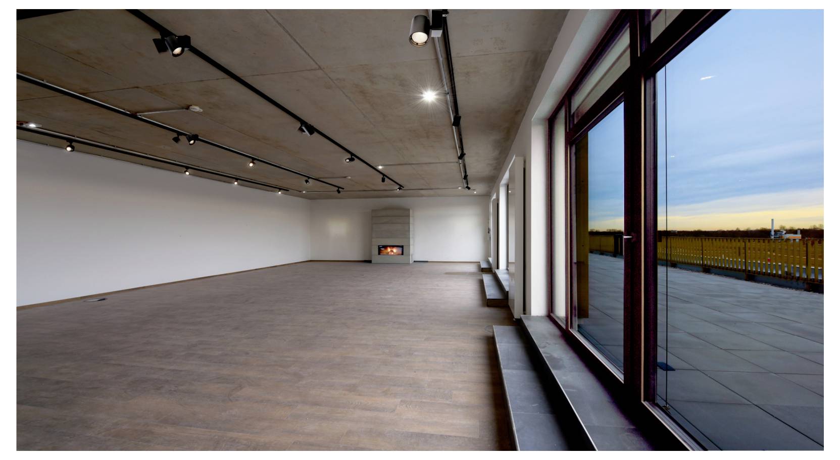 Pièce moderne vide avec parquet, éclairage sur rail et grandes baies vitrées du sol au plafond donnant sur une terrasse extérieure et un paysage.