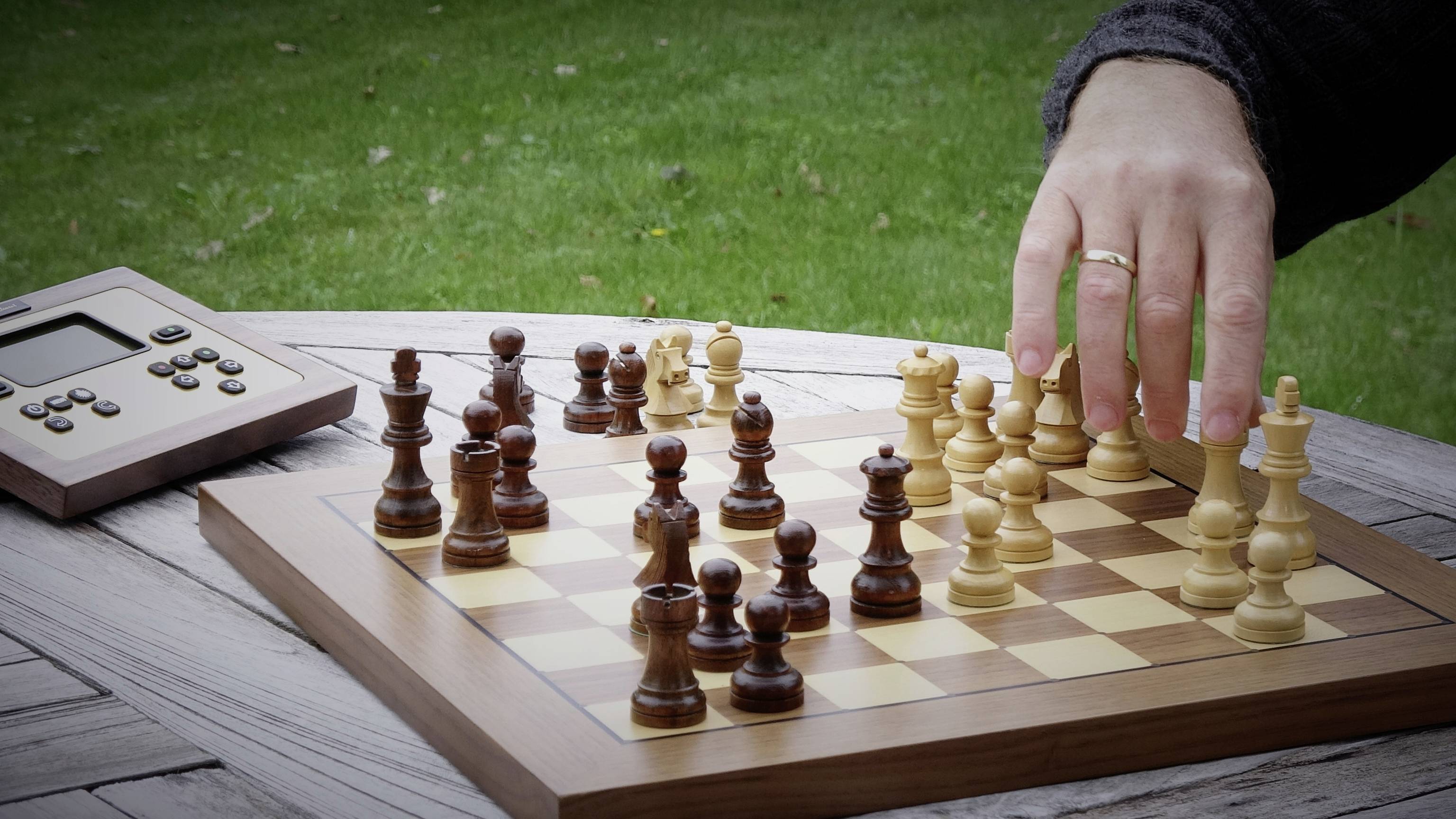 Une main déplace un pion blanc sur un échiquier. Un chronomètre d'échecs numérique est posé à côté sur une table en bois à l'extérieur.