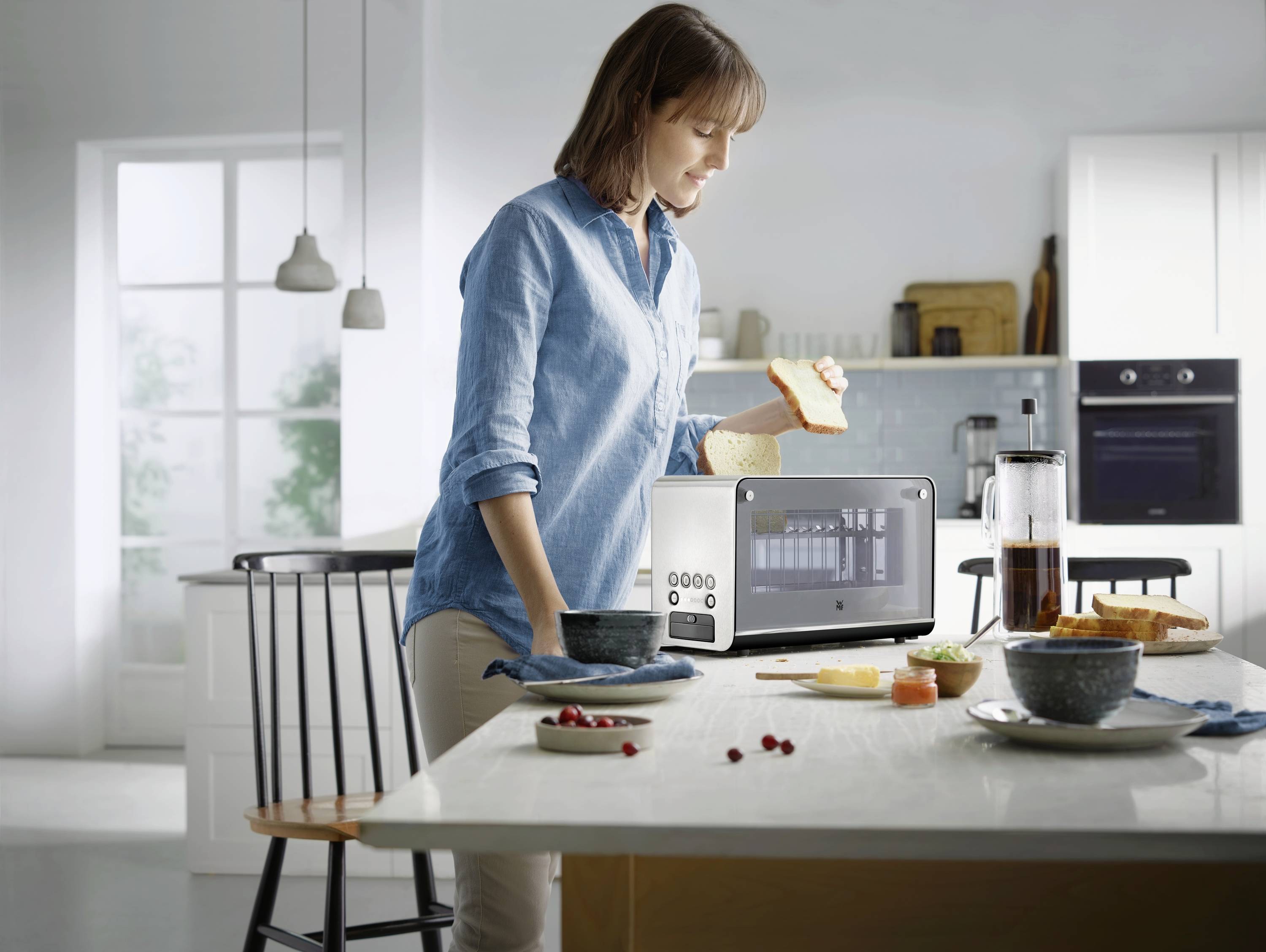 Une femme prépare du pain grillé dans une cuisine moderne. Sur la table se trouvent des bols, du pain et un grille-pain avec un affichage numérique.