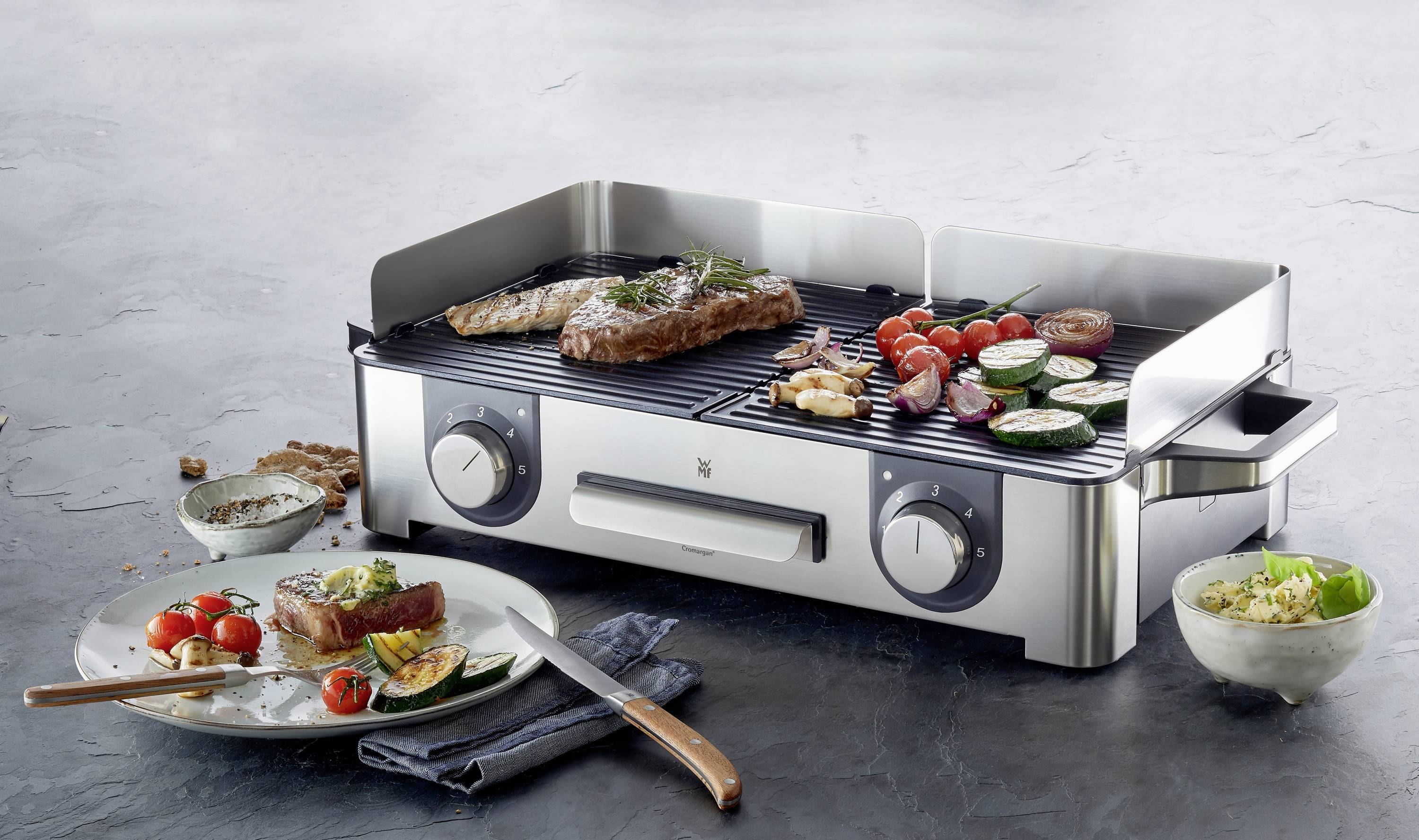 Un grill de table avec deux steaks, des tomates, des courgettes et des champignons dessus, entouré d'assiettes contenant des aliments grillés sur une table.