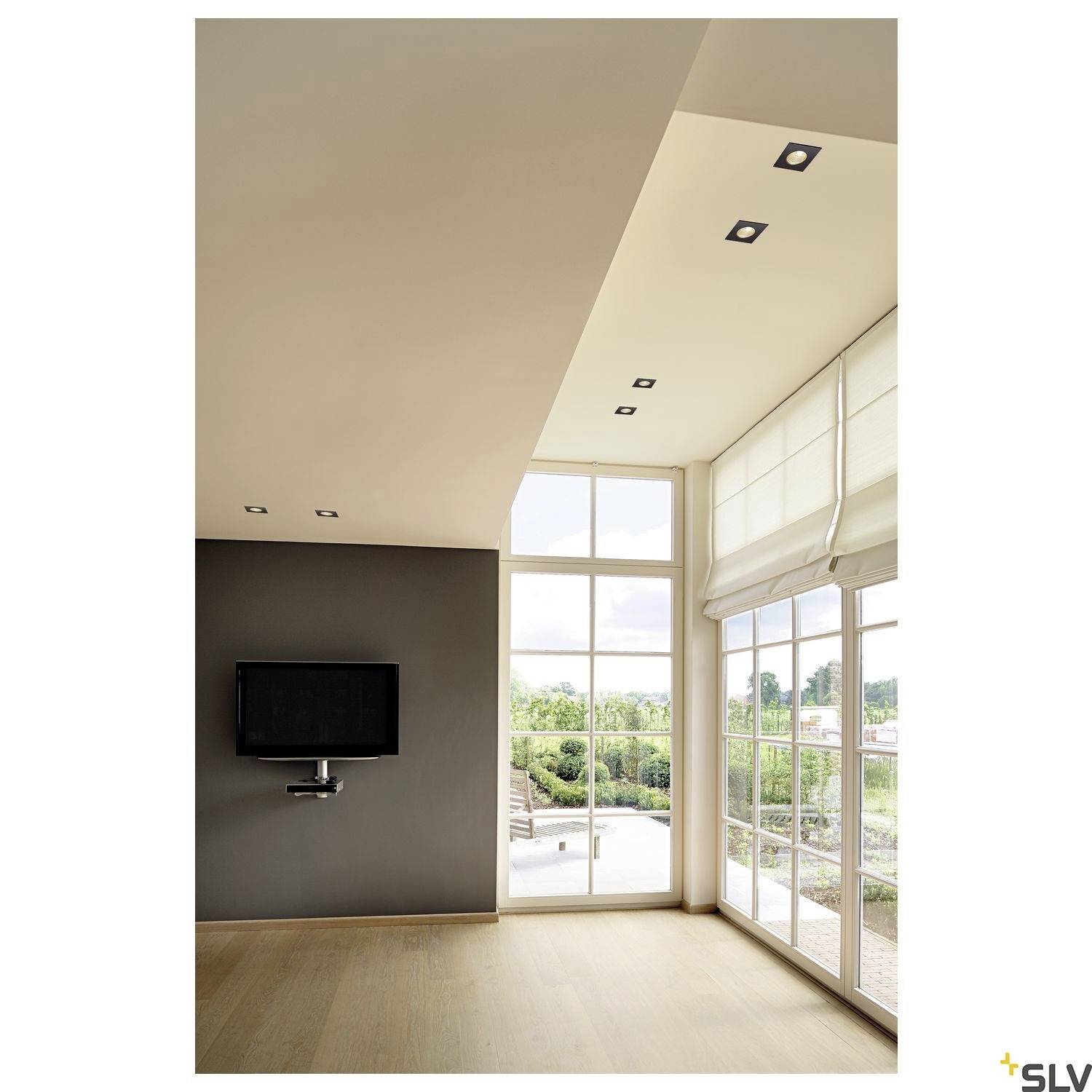 Salon avec une grande fenêtre et vue sur une terrasse. Éclairage de plafond moderne, avec un téléviseur à écran plat accroché au mur.