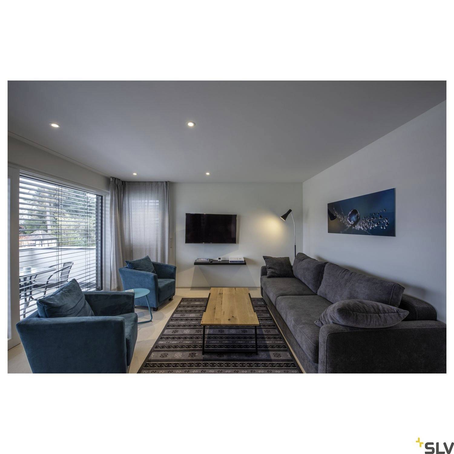 Espace de vie moderne avec un canapé gris, deux fauteuils bleus, une table en bois et une télévision au mur. À gauche, de grandes fenêtres avec des stores.
