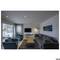 Espace de vie moderne avec un canapé gris, deux fauteuils bleus, une table en bois et une télévision au mur. À gauche, de grandes fenêtres avec des stores.