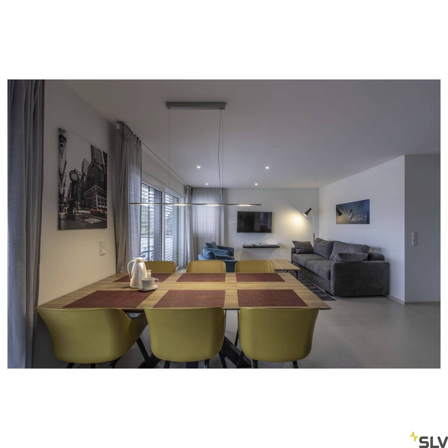 Salle à manger avec une grande table en bois et des chaises jaunes, donnant sur un salon moderne avec un canapé gris et une télévision murale, éclairage lumineux.