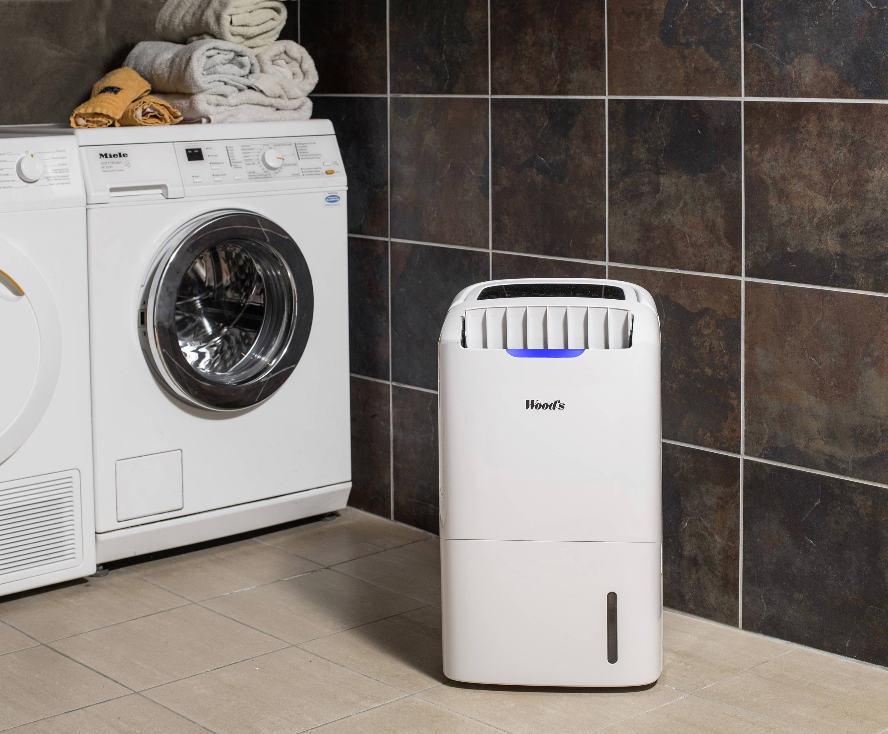 Un sèche-linge blanc et un déshumidificateur sont côte à côte devant un mur carrelé, avec des serviettes sur le sèche-linge.