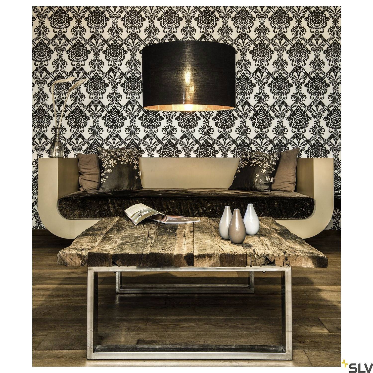 Un espace de vie élégant avec une lampe suspendue noire, un canapé à motifs contre un mur de papier peint ornemental et une table en bois rustique.