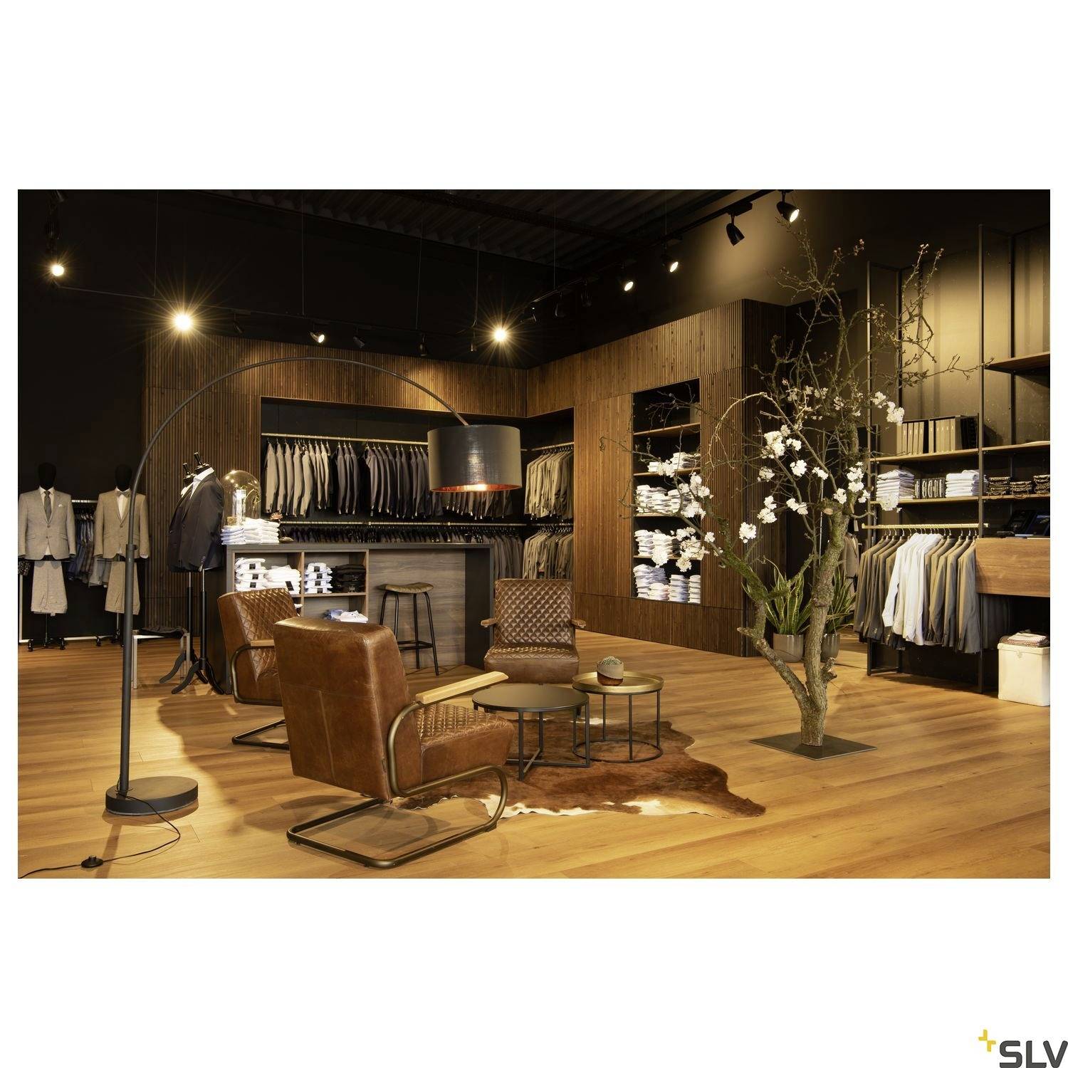 Boutique exclusive de vêtements avec des fauteuils en cuir, un sol en bois, des étagères remplies de vêtements et un concept d'éclairage moderne.