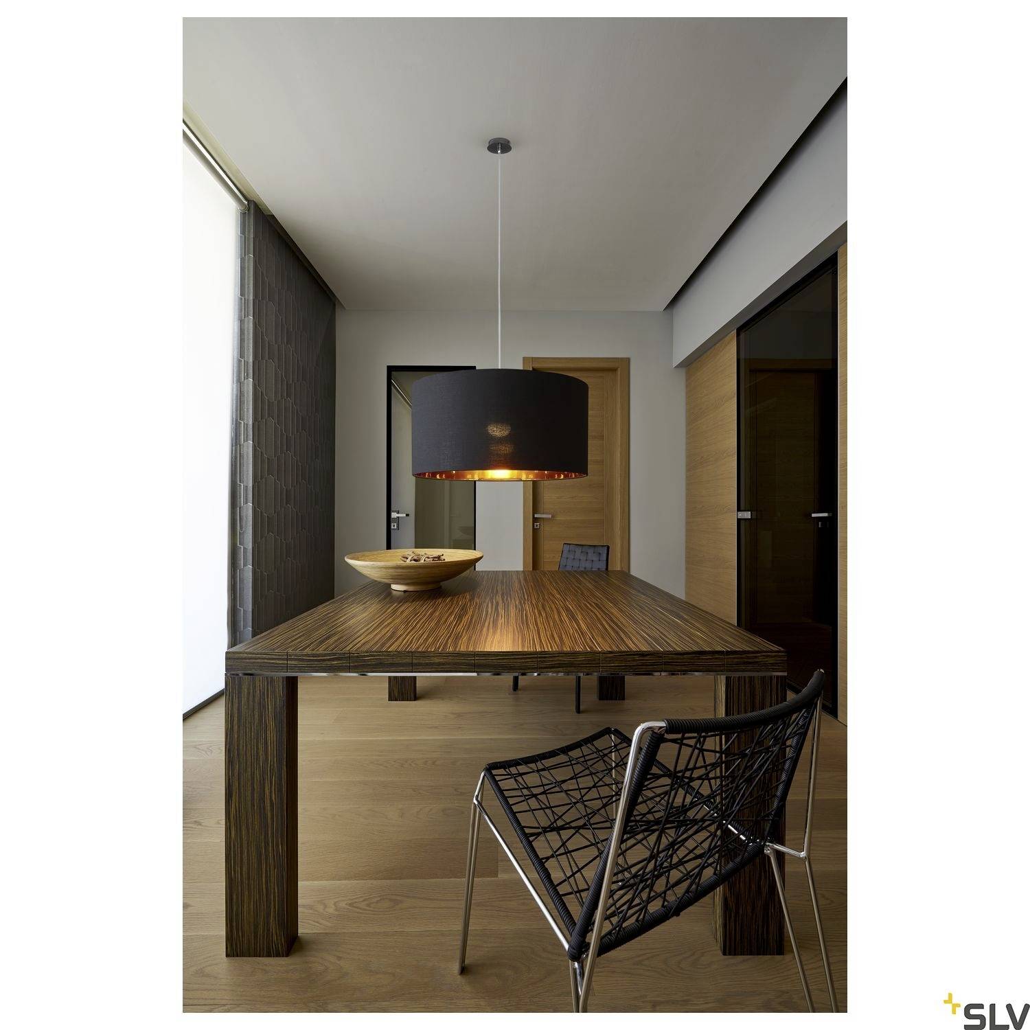 Un espace salle à manger moderne avec une table en bois, sur laquelle repose un bol en bois. Une suspension noire est suspendue au-dessus. À l'arrière-plan, une porte et des fenêtres.