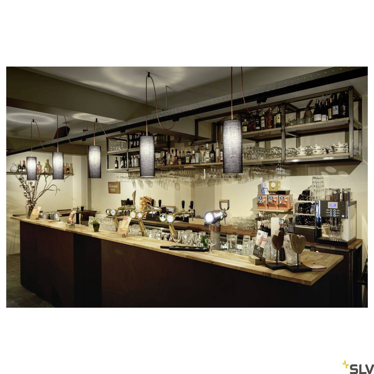 Bar avec comptoir en bois, éclairée par des lampes suspendues. En arrière-plan, des étagères remplies de bouteilles et de verres. À gauche, une décoration avec des branches.