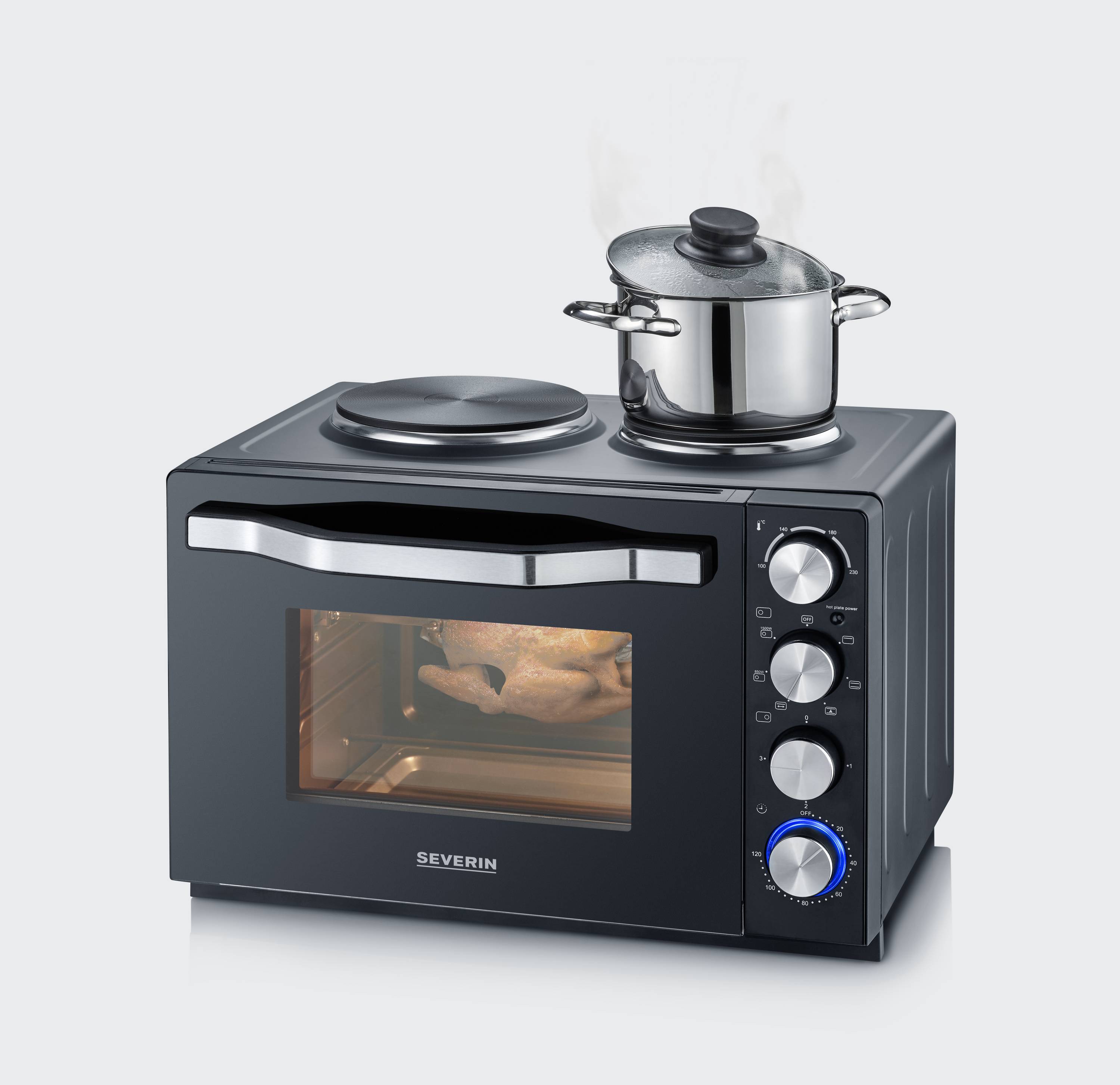 Four compact avec deux plaques de cuisson sur le dessus, une casserole avec de la vapeur est posée dessus. À l'intérieur du four, on peut voir un poulet rôti.