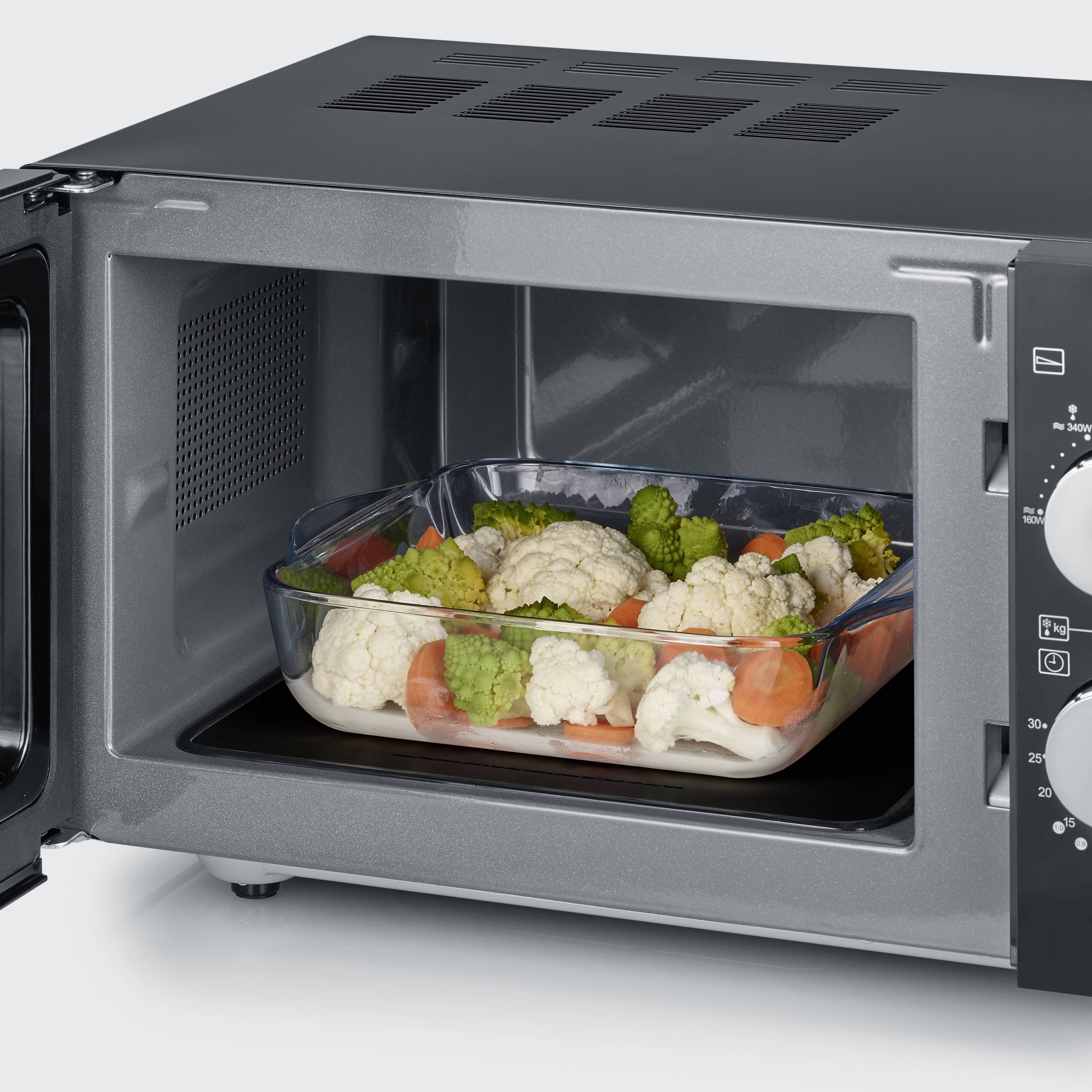 Un four à micro-ondes ouvert avec un récipient en verre contenant des légumes tels que du chou-fleur et des carottes. Le micro-ondes est posé sur un plan de travail.