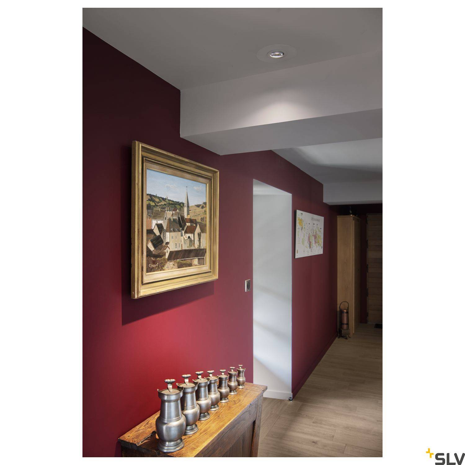 Un couloir rouge avec une table en bois au premier plan et six vases en métal posés dessus. Un tableau de paysage est accroché au mur.