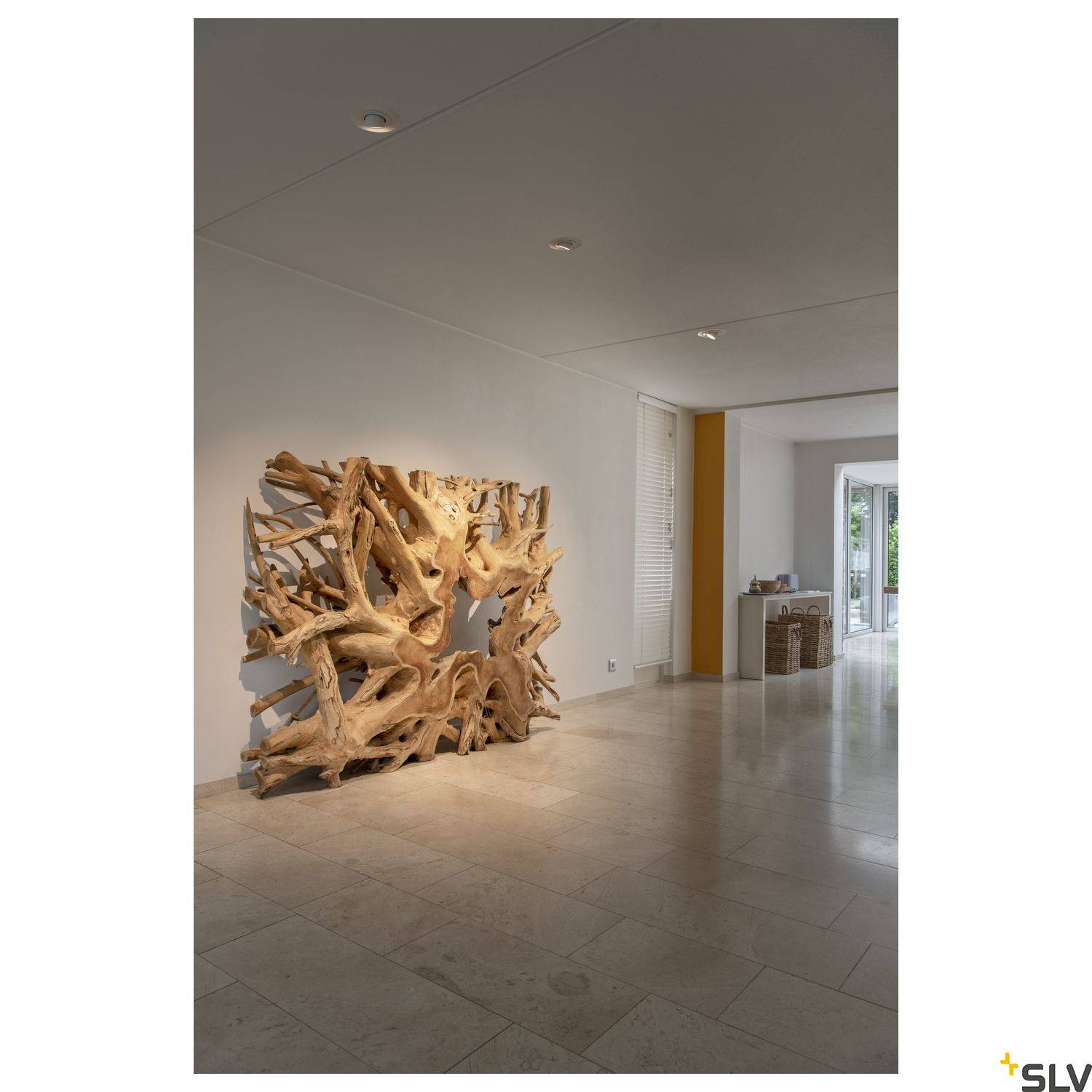 Une grande œuvre d'art organique en bois est exposée contre un mur blanc dans une galerie lumineuse. À droite, un passage mène à une autre pièce baignée de lumière naturelle.