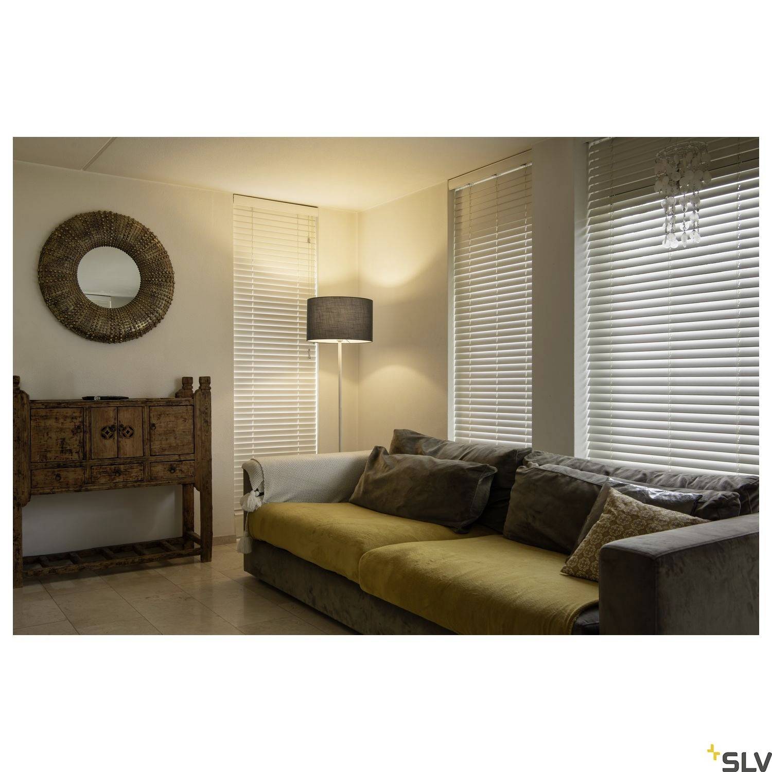 Salon avec un canapé gris, plusieurs coussins, des meubles en bois avec un miroir rond et un lampadaire dans un éclairage tamisé.