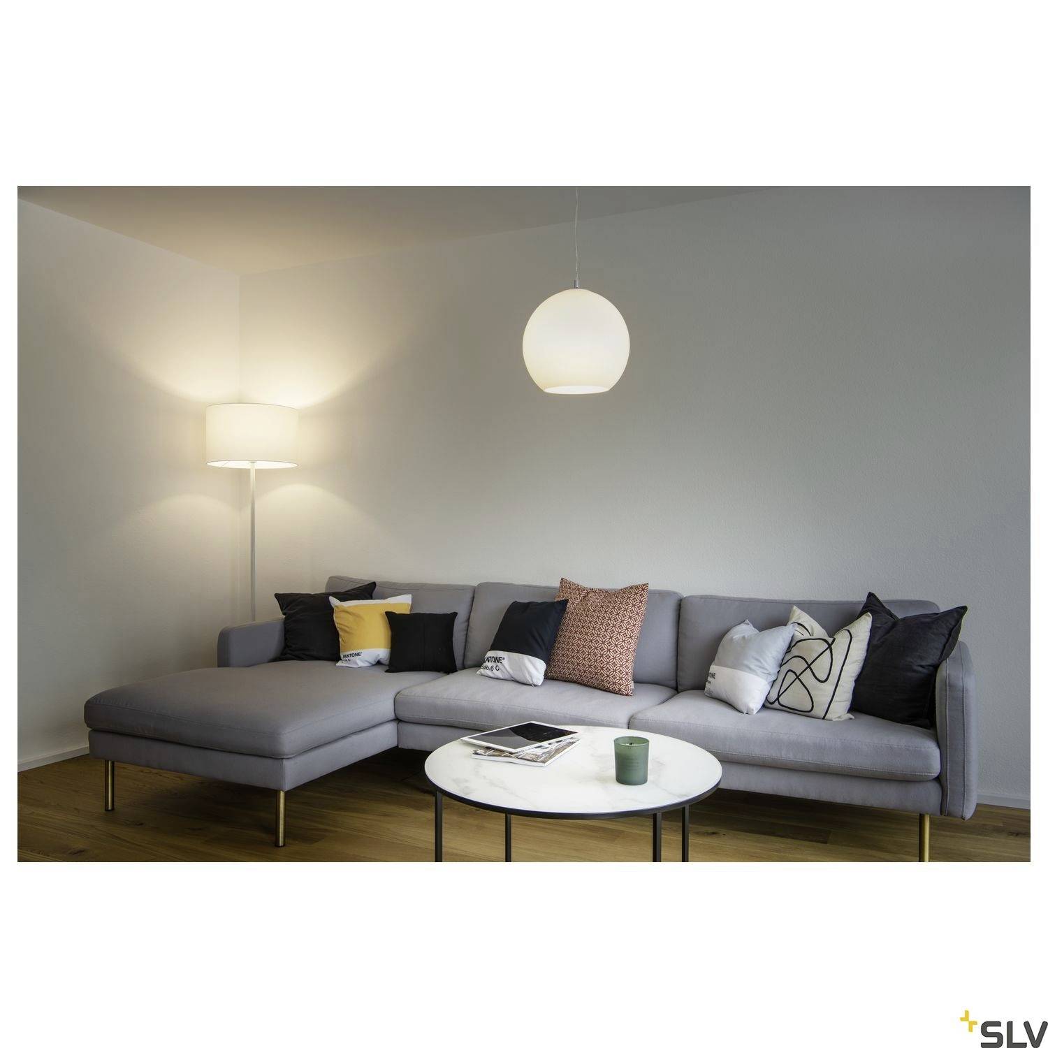 Un salon chaleureux avec un canapé gris, plusieurs coussins et une table basse ronde. Un lampadaire et une suspension éclairent la pièce.