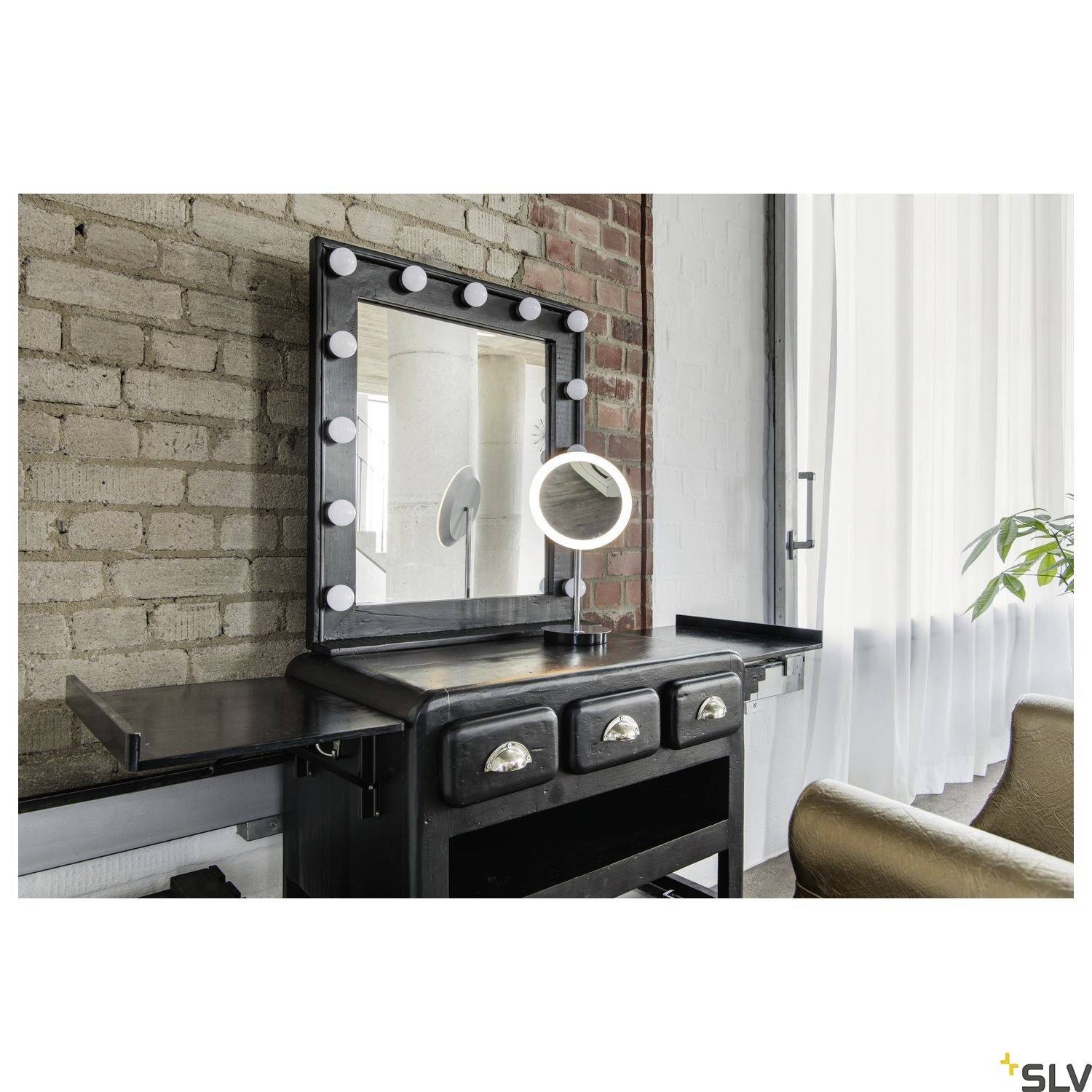 Coiffeuse avec miroir éclairé devant un mur de briques, à côté une chaise marron et une fenêtre avec un rideau blanc.