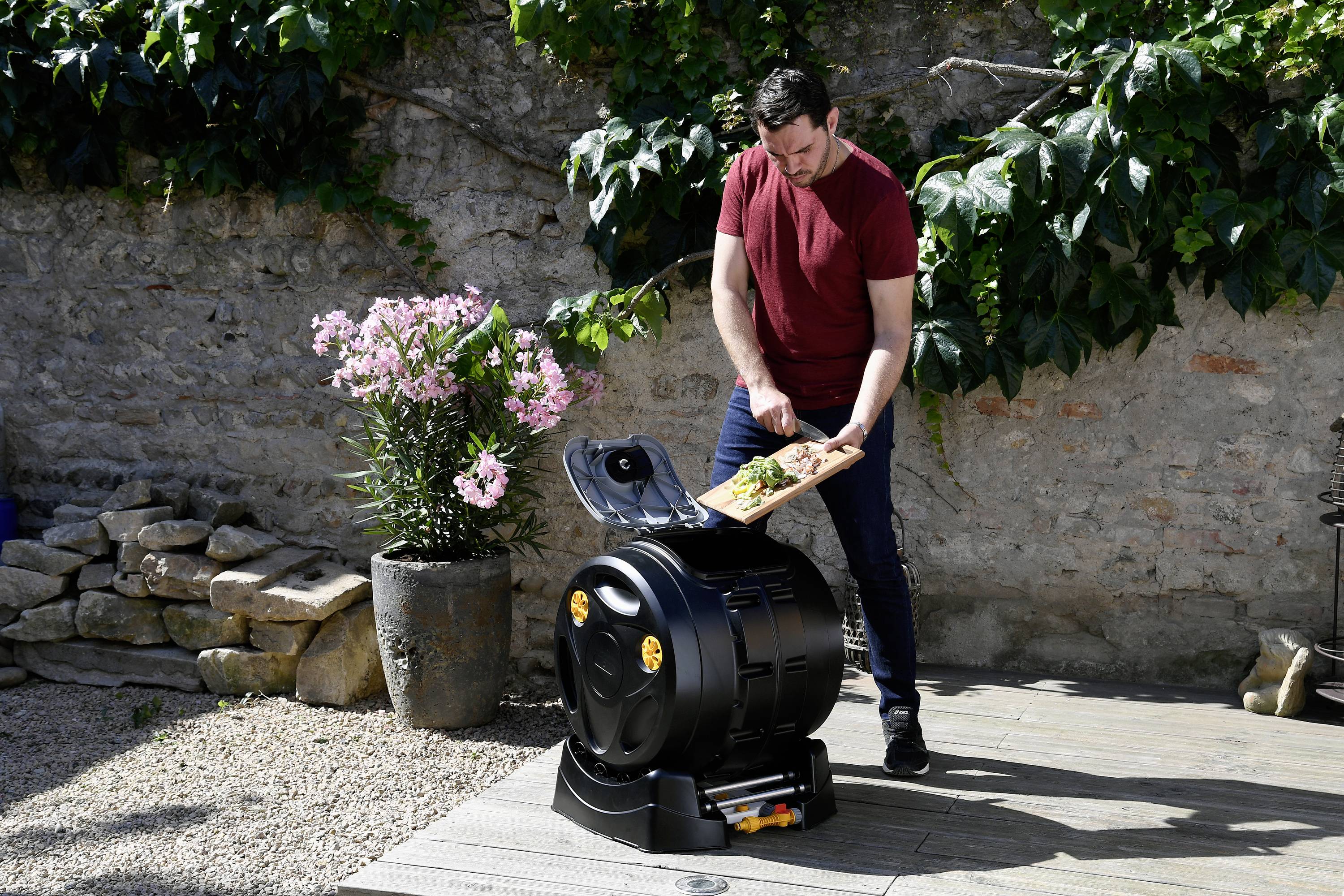 Un homme remplit un composteur avec des déchets organiques dans un jardin. À côté de lui se trouvent des plantes en fleurs, et un mur de pierre est visible en arrière-plan.