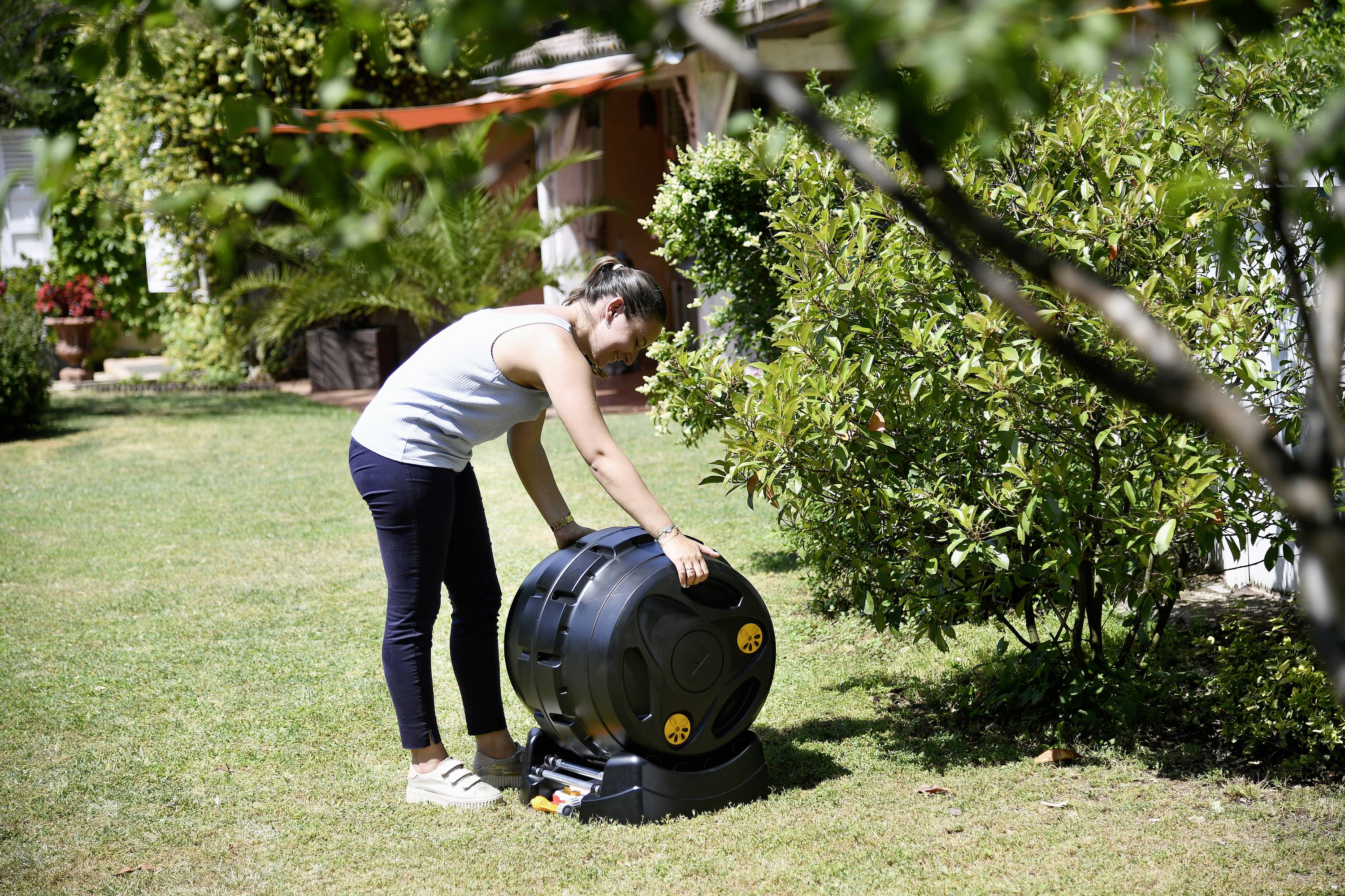 Une femme fait tourner un composteur dans un jardin. Des arbres et des arbustes sont visibles en arrière-plan.
